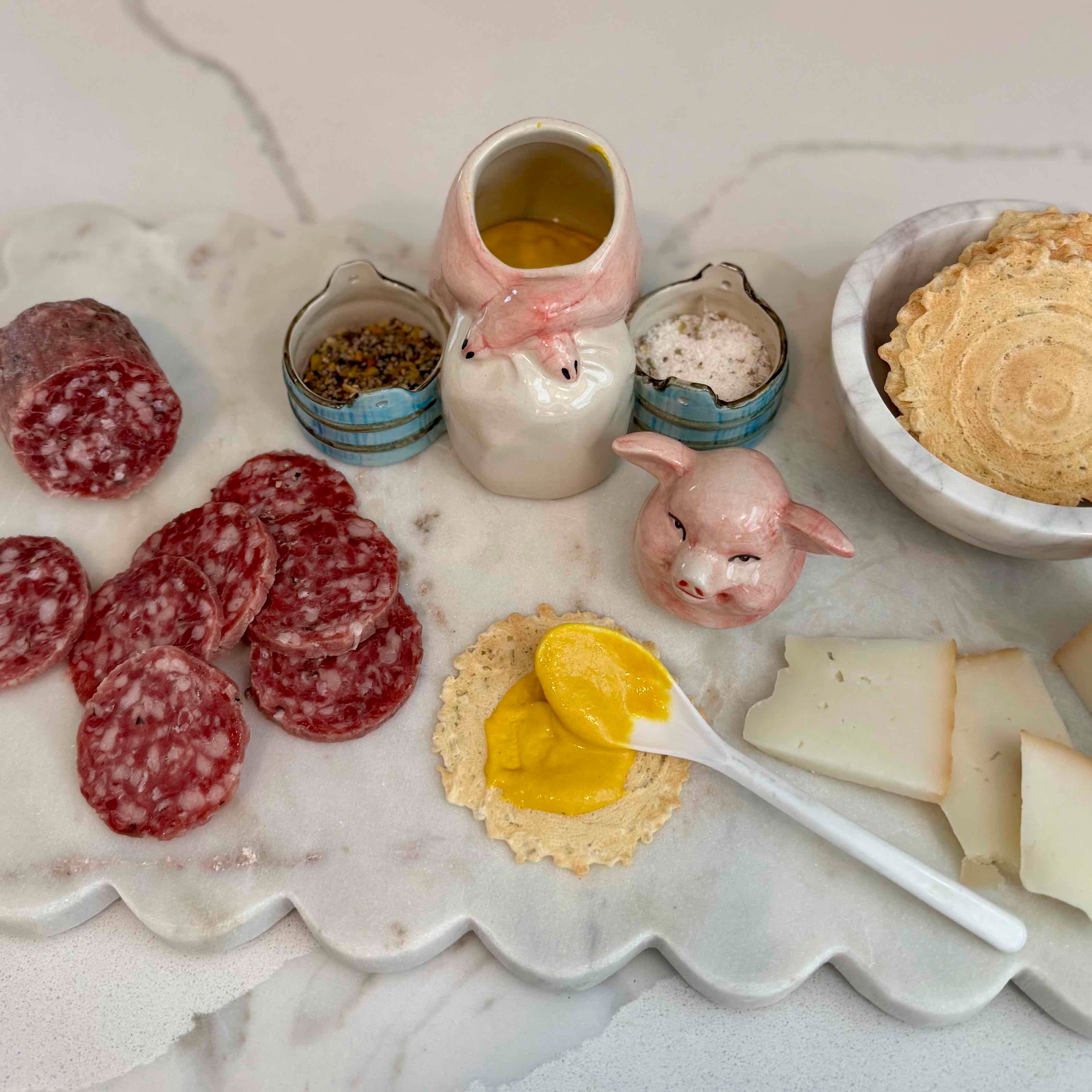 Platter with sliced salami, crackers, and a small container of yellow spread on a marble surface.