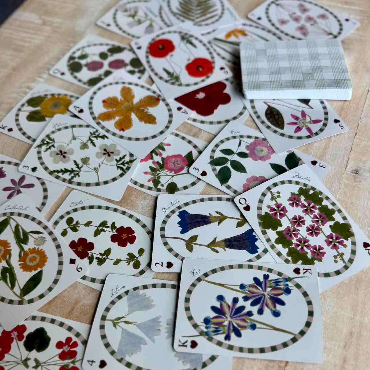 A spread of illustrated playing cards on a light wood surface, with a small stack of cards showing a white-and-green plaid back. The cards feature oval floral and botanical designs in many colors including red, pink, blue, yellow, green, and white.