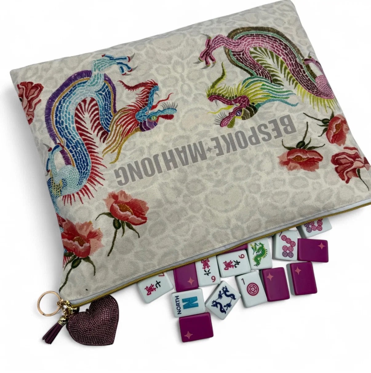 A decorative zippered pouch with a beige patterned background, embroidered phoenix motifs in blue, pink, and green, and red rose illustrations. The pouch is shown open with several miniature mahjong tiles arranged in front, and a dark purple heart-shaped tassel keychain hangs from the zipper.