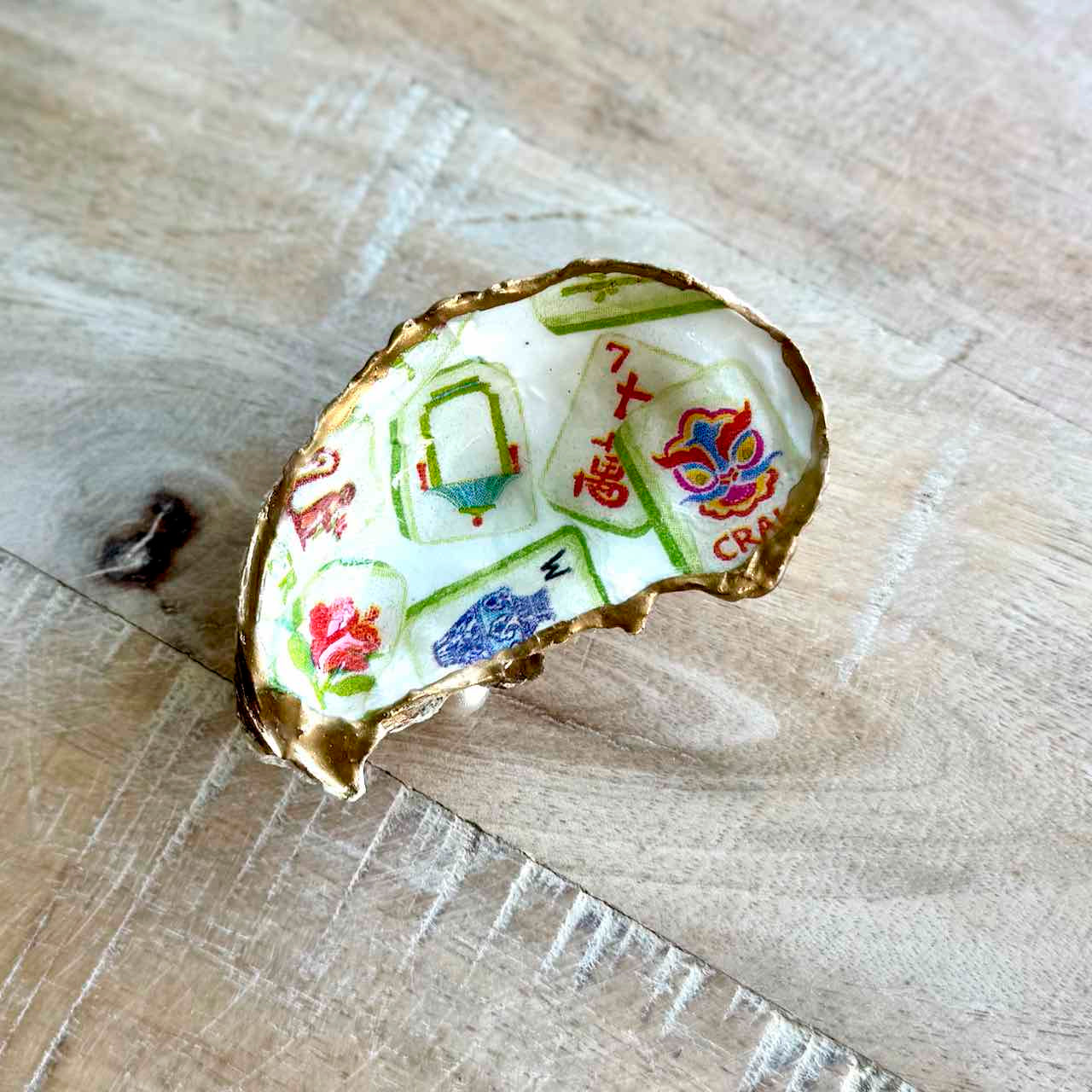 A handmade ring shaped like a broken shell, featuring a white enamel face decorated with colorful Chinese-style tiles, characters, and motifs, and finished with a gold-tone rim, resting on a light wood surface.