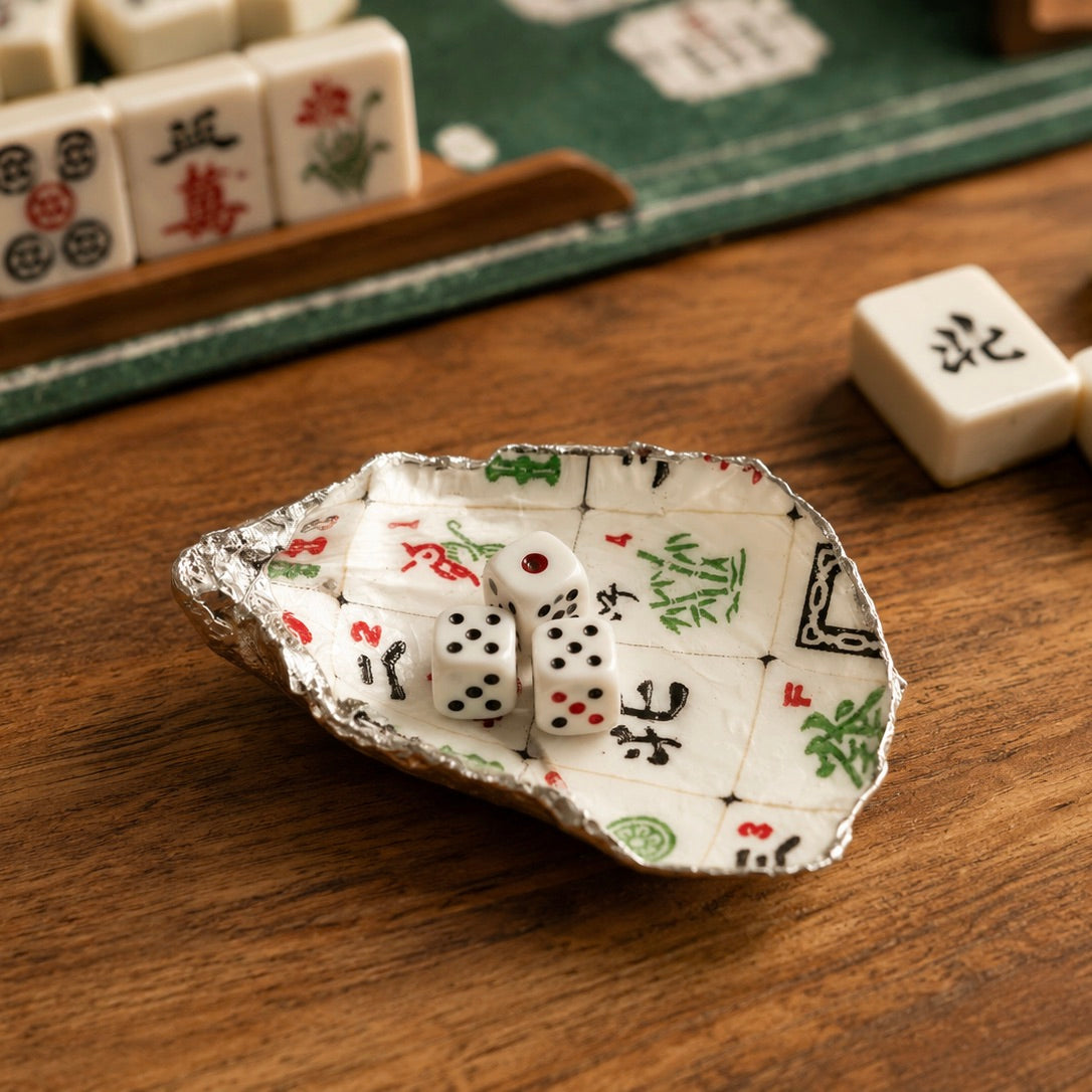 A small decorative dish shaped like an open shell, printed with a mahjong board design and holding two white dice, set on a wooden tabletop with mahjong tiles nearby.