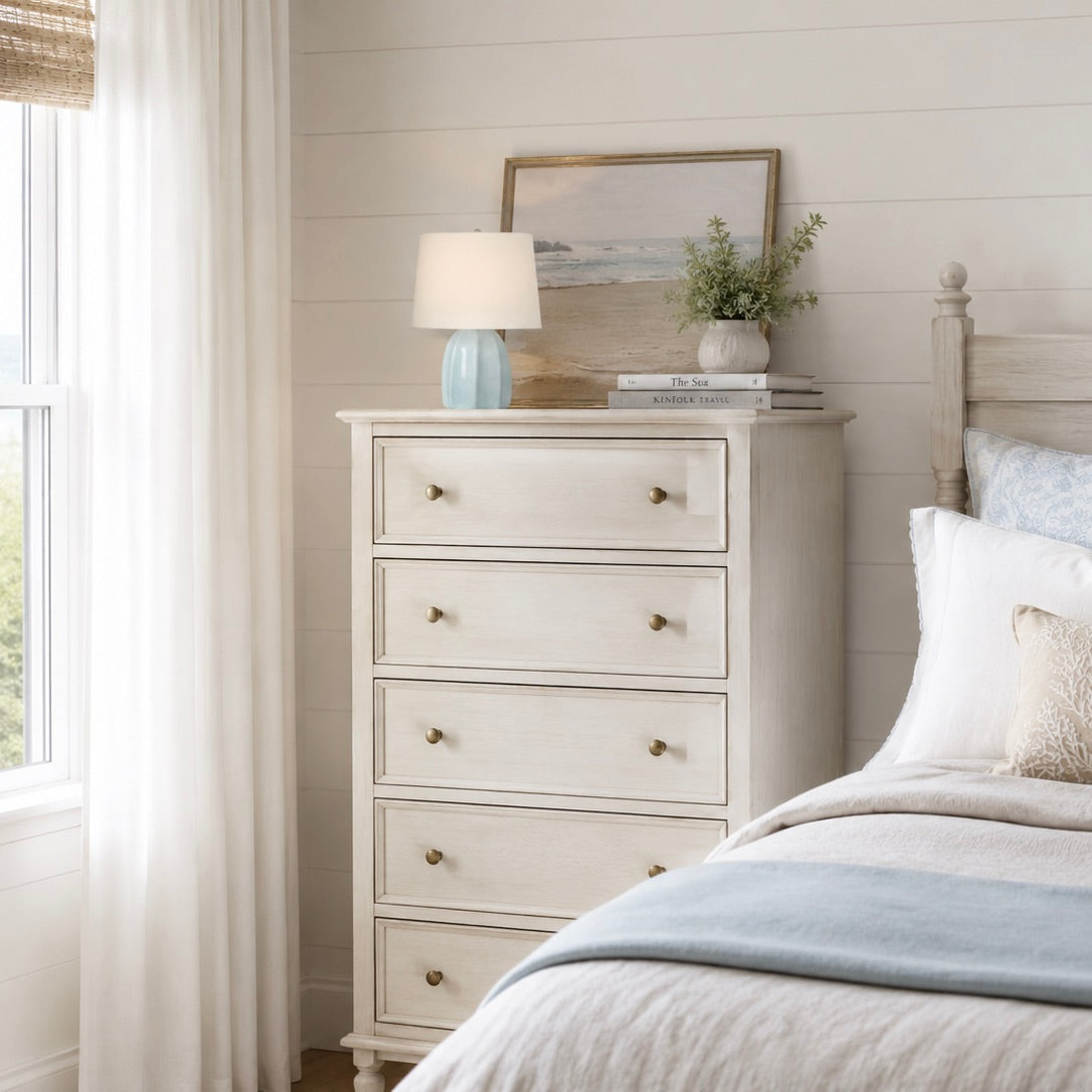 An off-white five-drawer dresser with brass knobs in a bright, neutral bedroom setting next to a bed with white and blue bedding.