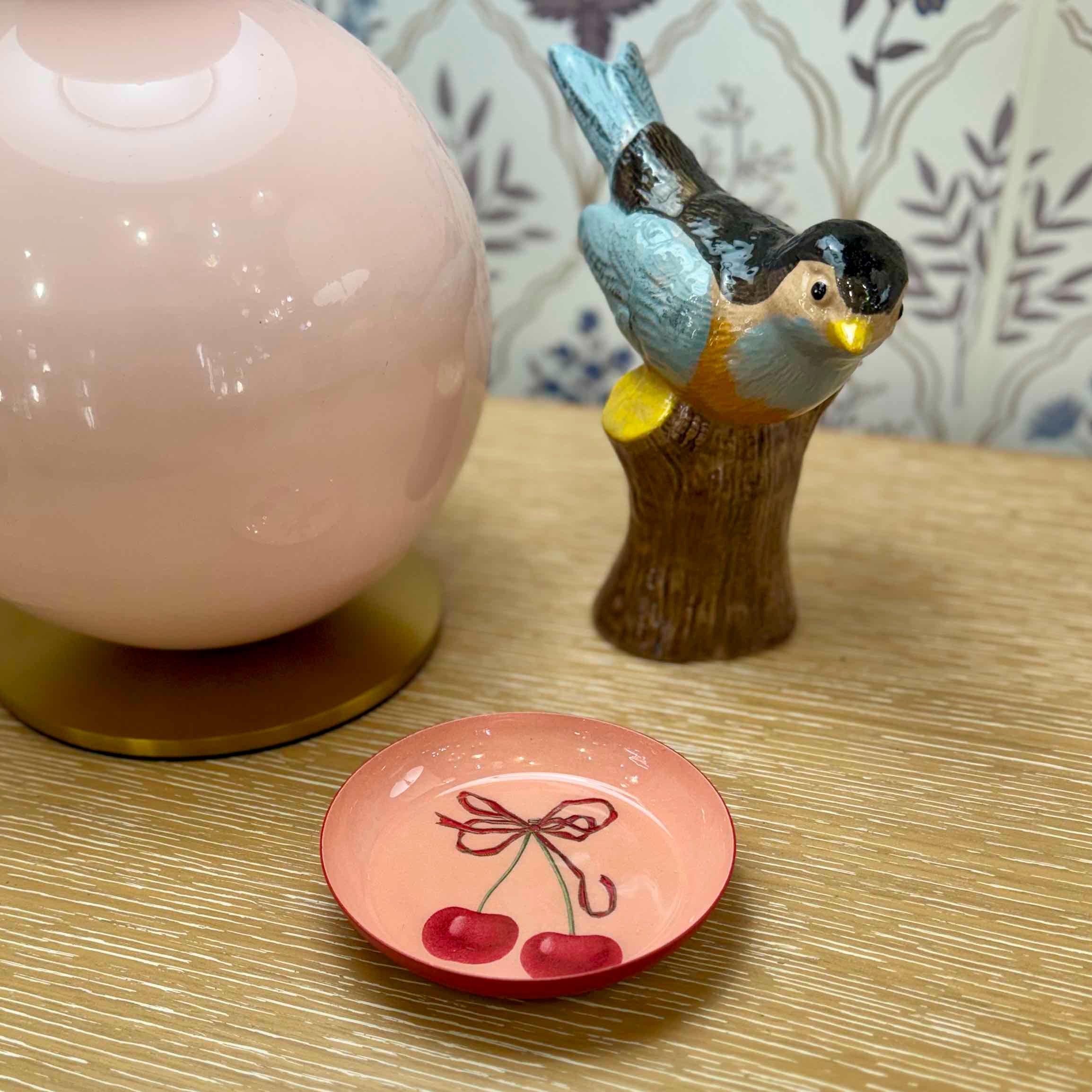 Decorative ceramic bird on a branch and a small pink plate with cherry design on a wooden surface.