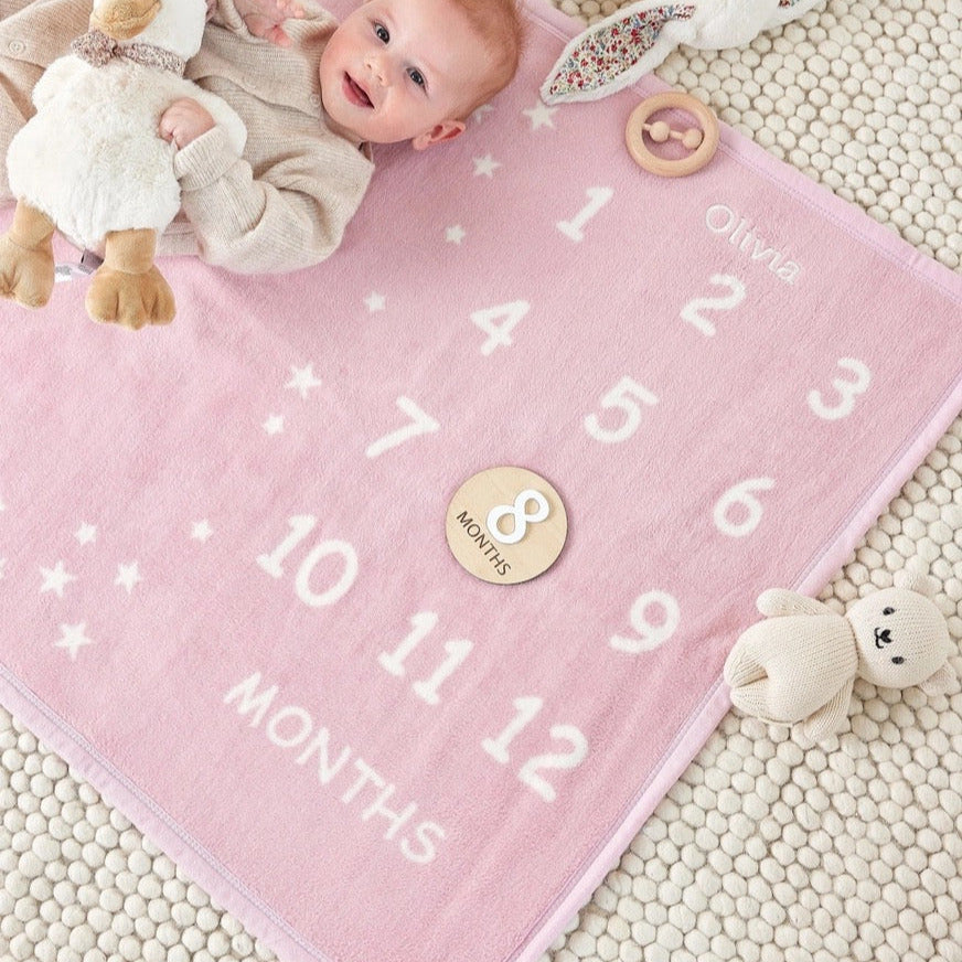 Pink milestone blanket with baby toys and a baby lying on it