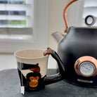 Black teapot with a monkey design on the spout and a white mug with a tea bag inside, on a surface.