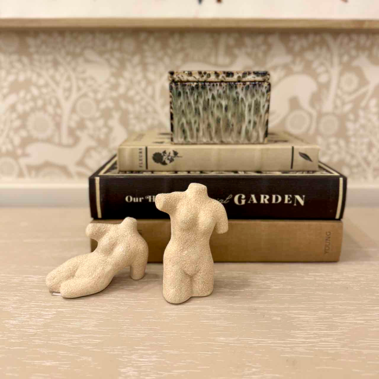 Two small beige sculptures of human figures on a textured surface with a book and decorative pot in the background.