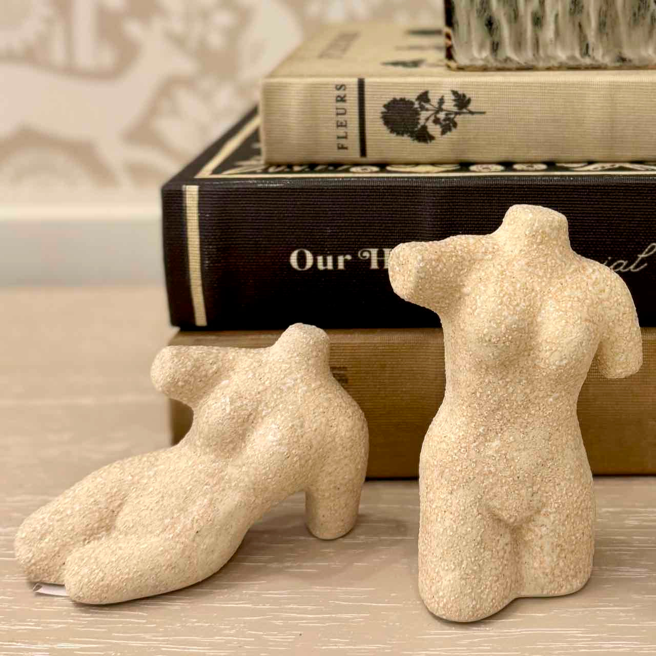 Decorative sand-textured torso and foot sculptures on a surface with books in the background.