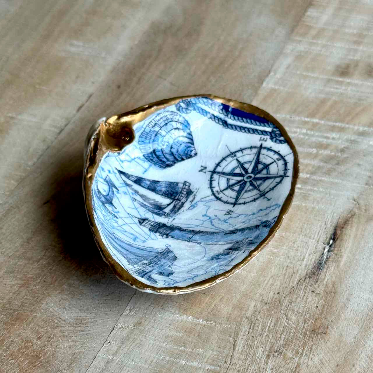 A small decorative shell-shaped dish with a blue and white nautical print of ships, waves, and a compass rose, finished with a gold-colored rim, resting on a light wood surface.