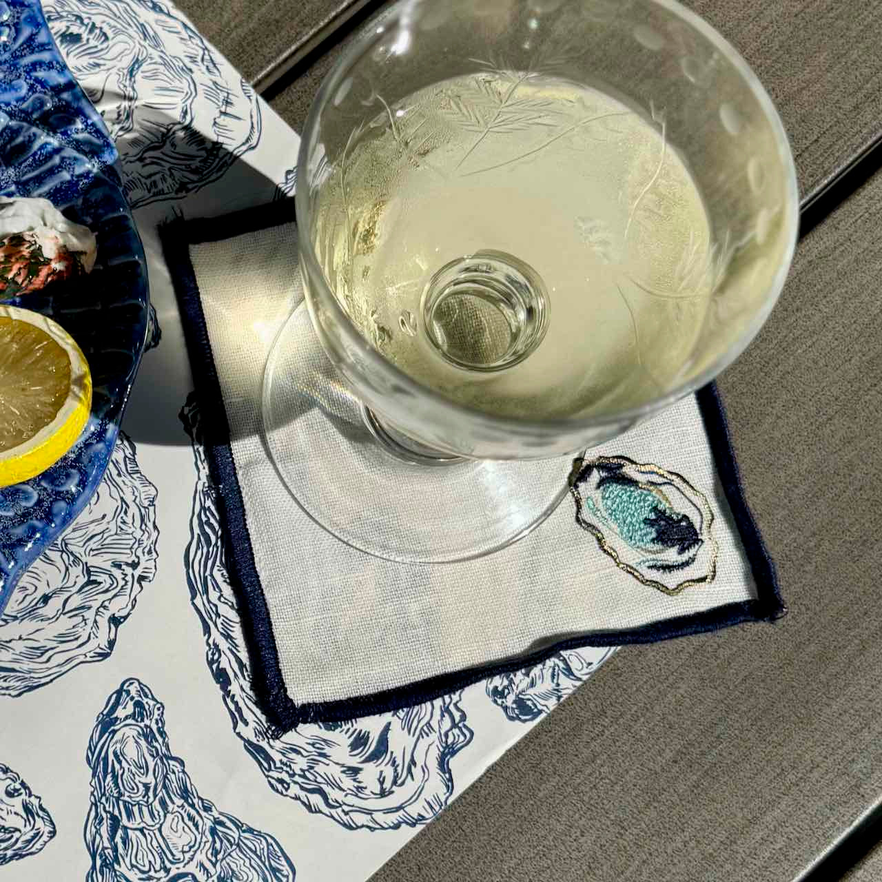 A clear stemmed glass containing a pale drink, placed on a white napkin with a navy embroidered oyster motif. The napkin rests on a gray table surface, with part of a blue-and-white patterned plate and a lemon slice visible at the left edge.