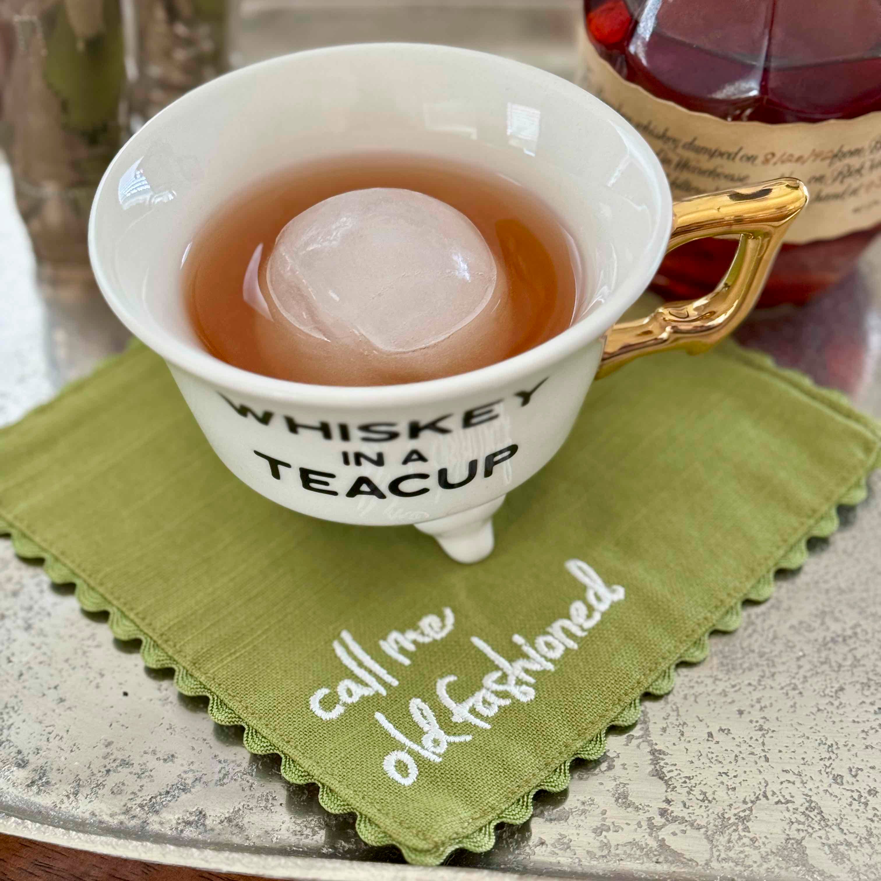 White teacup with gold handle containing a round ice cube in a liquid, on a green napkin with text.