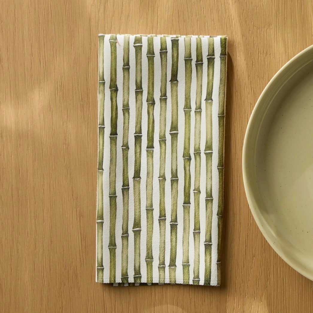 A rectangular woven placemat with a vertical bamboo-style pattern in olive green and off-white, laid flat on a light wood tabletop next to a ceramic plate.