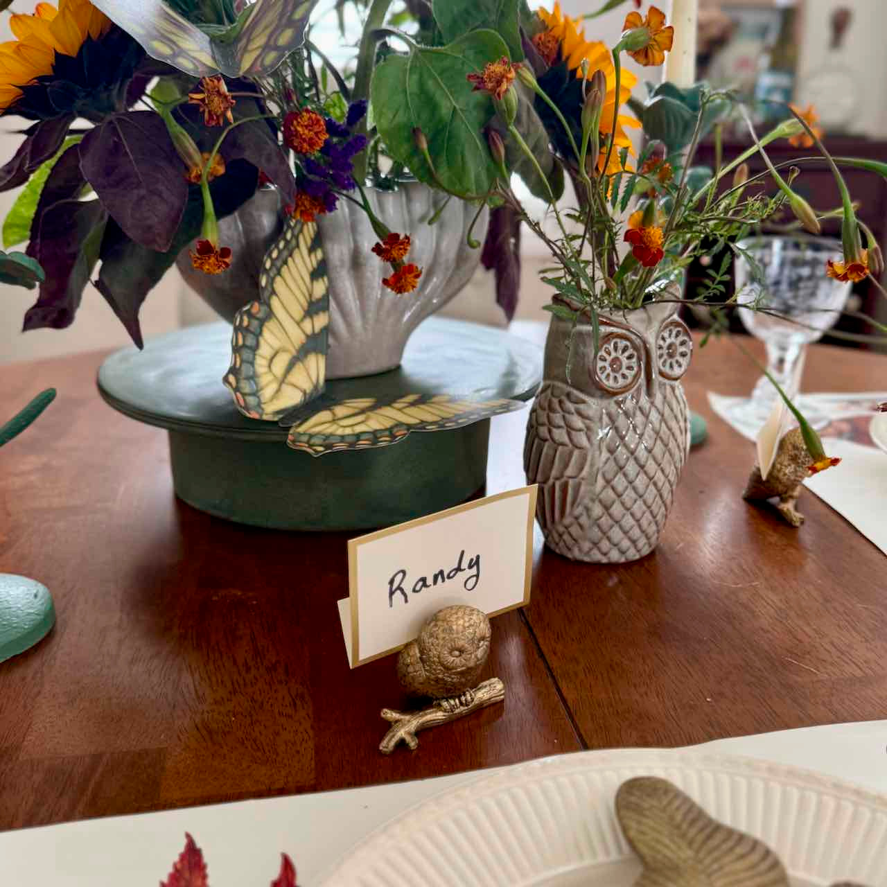 Decorative table setting with flowers, butterfly, owl vase, and name card on a wooden table.