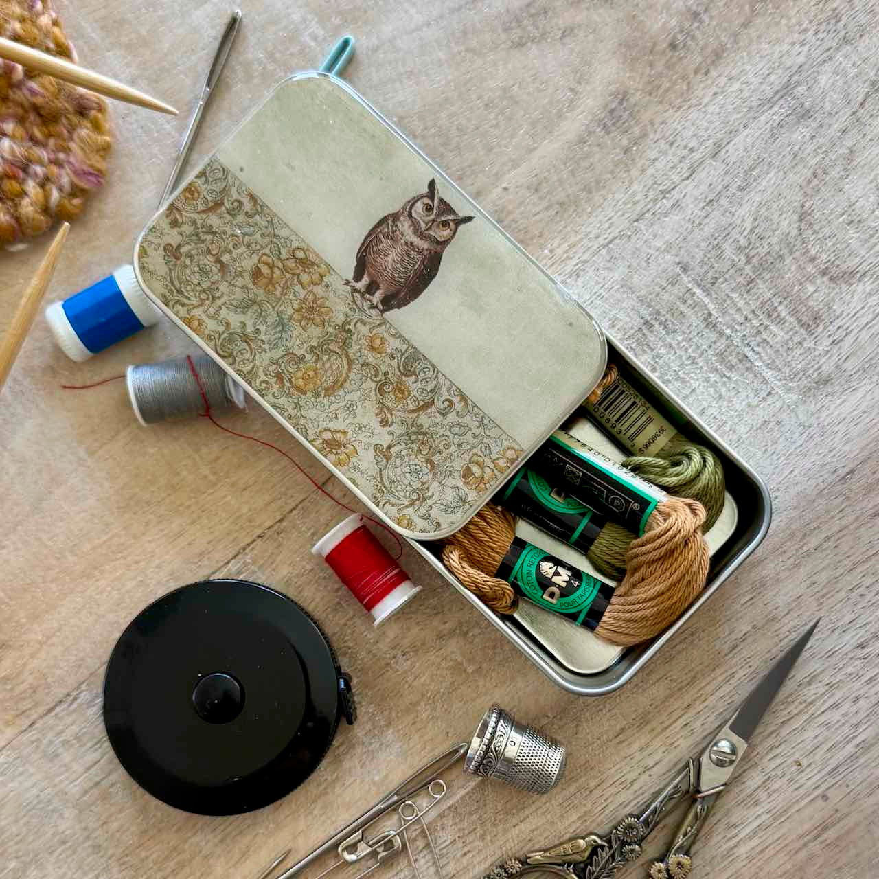 A metal sewing kit tin with a decorative owl illustration on the lid, shown open on a wooden surface with thread spools, scissors, a thimble, and other sewing tools arranged around it.