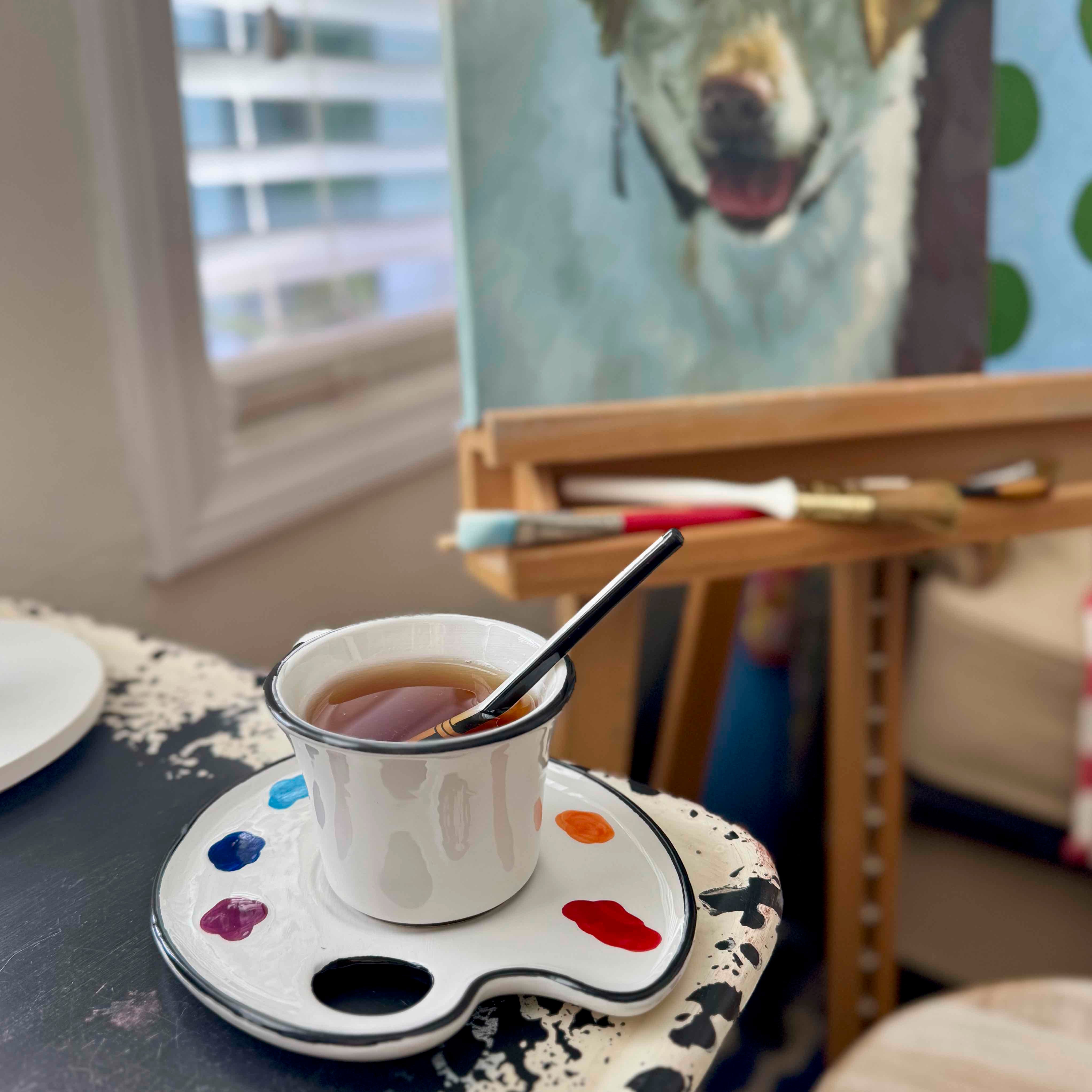 Cup of tea with a spoon on a paint palette in front of a painting of a dog.