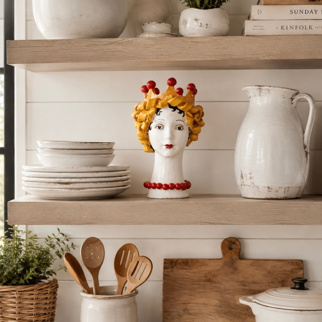 A decorative ceramic head sculpture with a yellow crown and red beads displayed on a wooden shelf in a kitchen setting, surrounded by white dishes, a white pitcher, and wooden utensils.