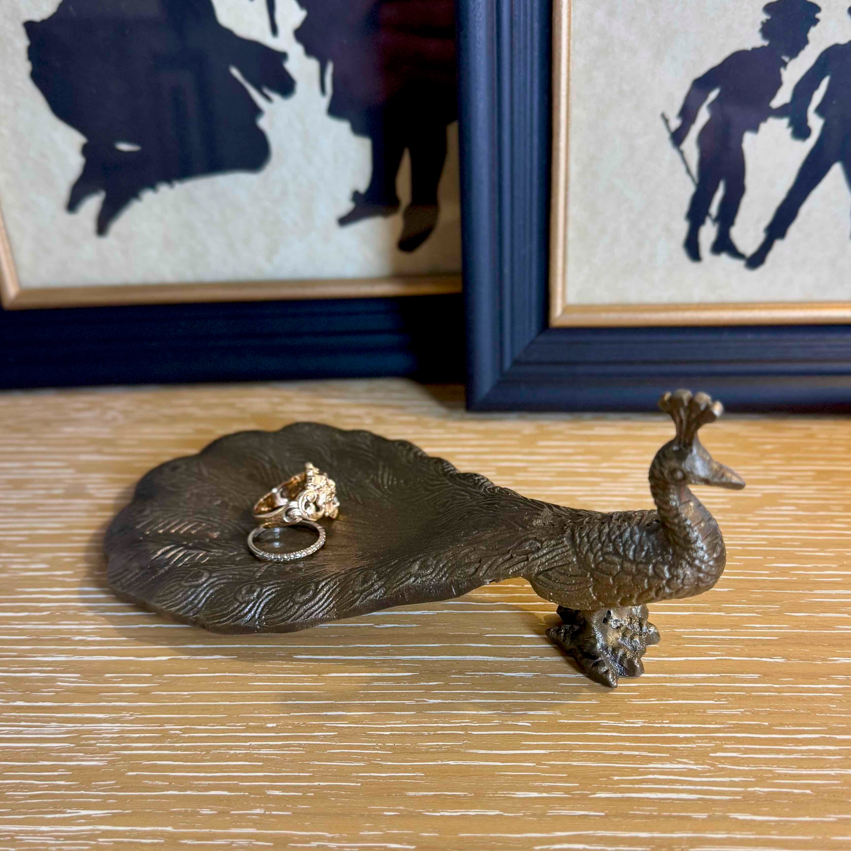 Bronze peacock-shaped ring holder with rings on a wooden surface, framed artwork in the background.