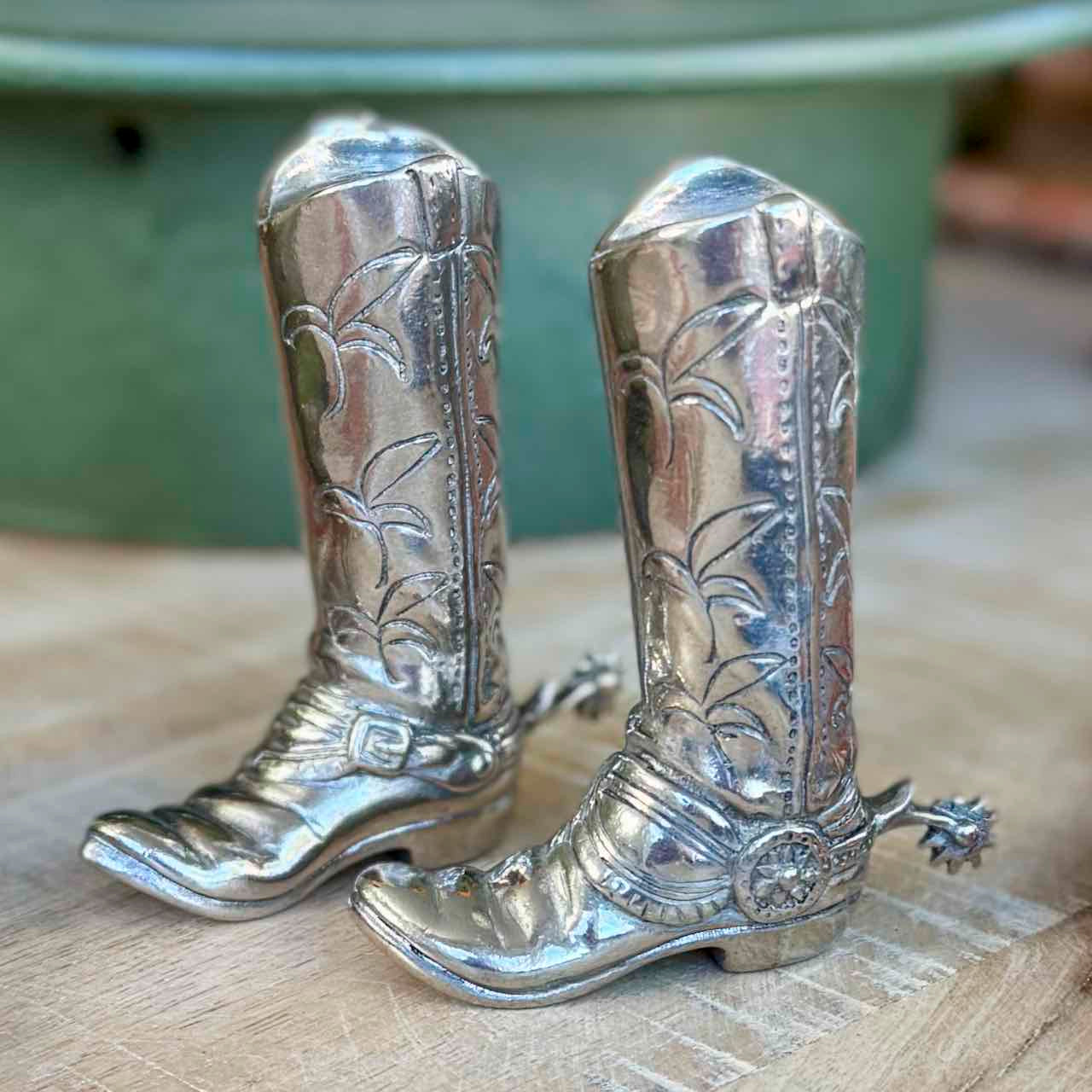 Silver cowboy boot salt and pepper shakers on a wooden surface with a blurred green background.