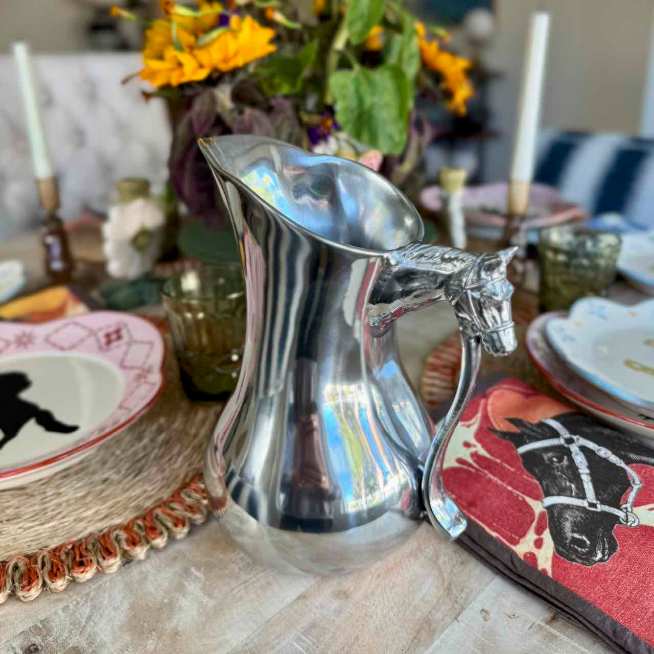 Silver pitcher with horse head design on a table with decorative plates and flowers.