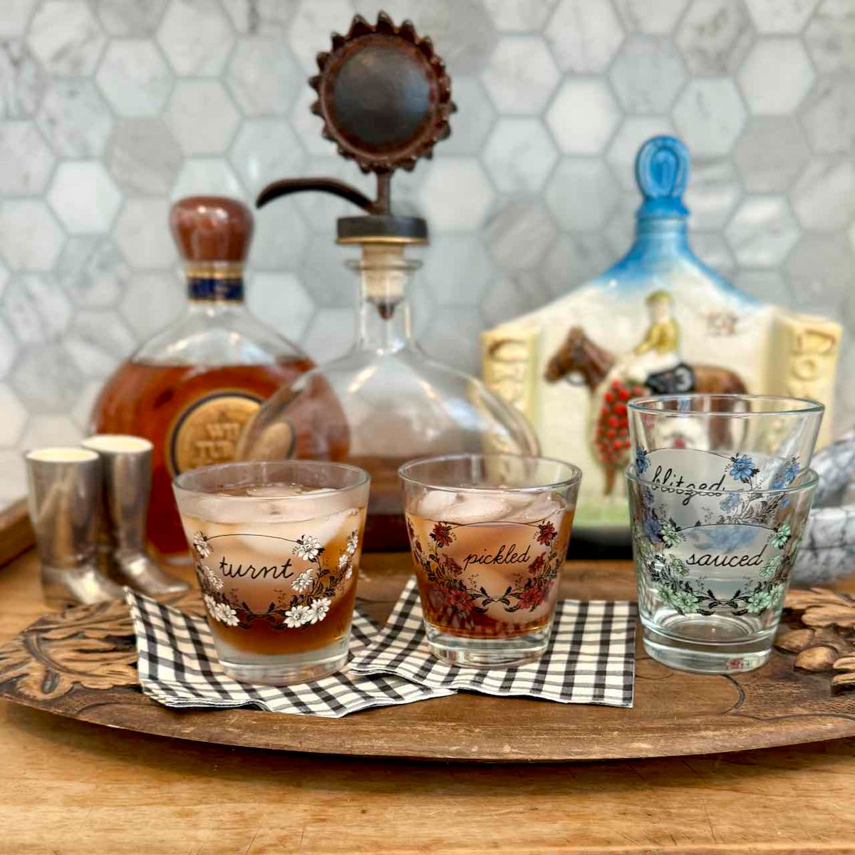 Three decorated clear drinking glasses with floral and script designs sit on a wooden tray in a kitchen setting, with bottles and a ceramic pitcher in the background.