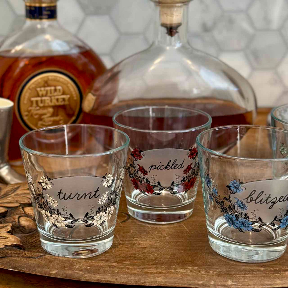 Three clear glass rocks tumblers with floral illustrations and handwritten labels are arranged on a wooden tray, with whiskey bottles in the blurred background.