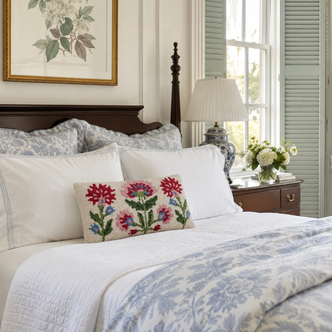 A styled bedroom scene with a bed dressed in white and light blue patterned bedding, featuring a decorative rectangular throw pillow with red and blue floral embroidery. The room includes a dark wood headboard, nightstand, lamp, window with shutters, and framed wall art.