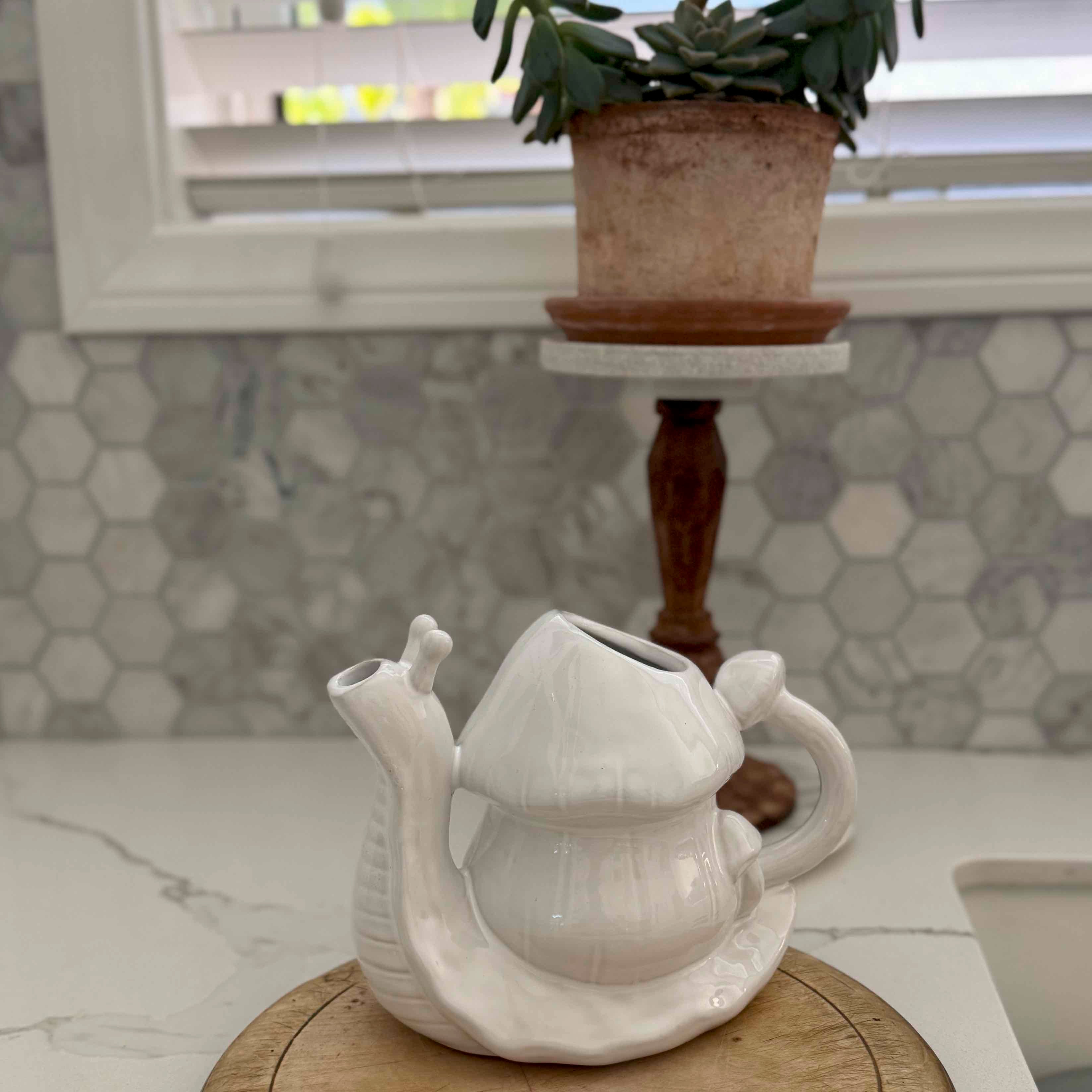 Snail-shaped ceramic teapot on a marble surface with a plant in the background.