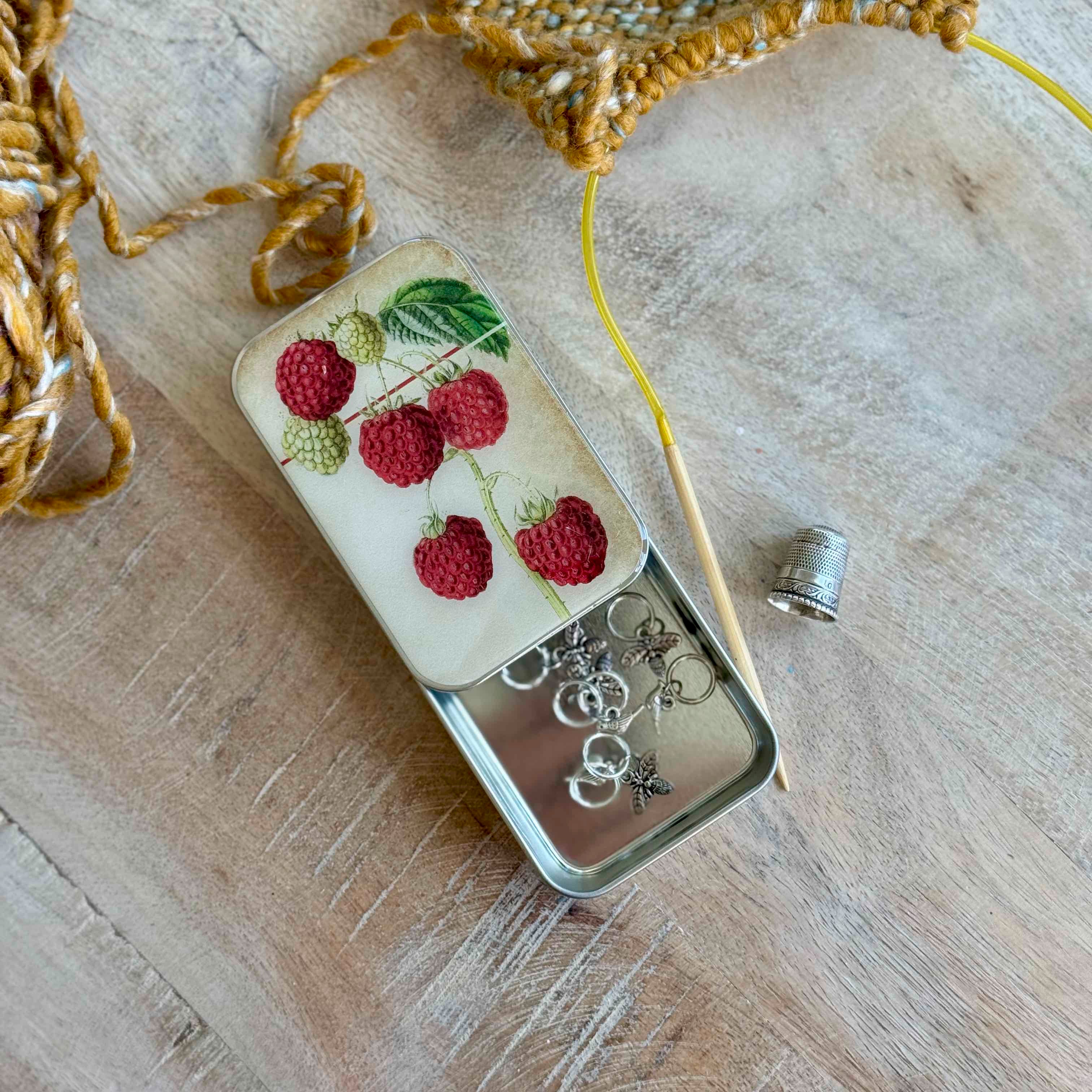 Small tin with raspberry design on lid, containing jewelry on a wooden surface