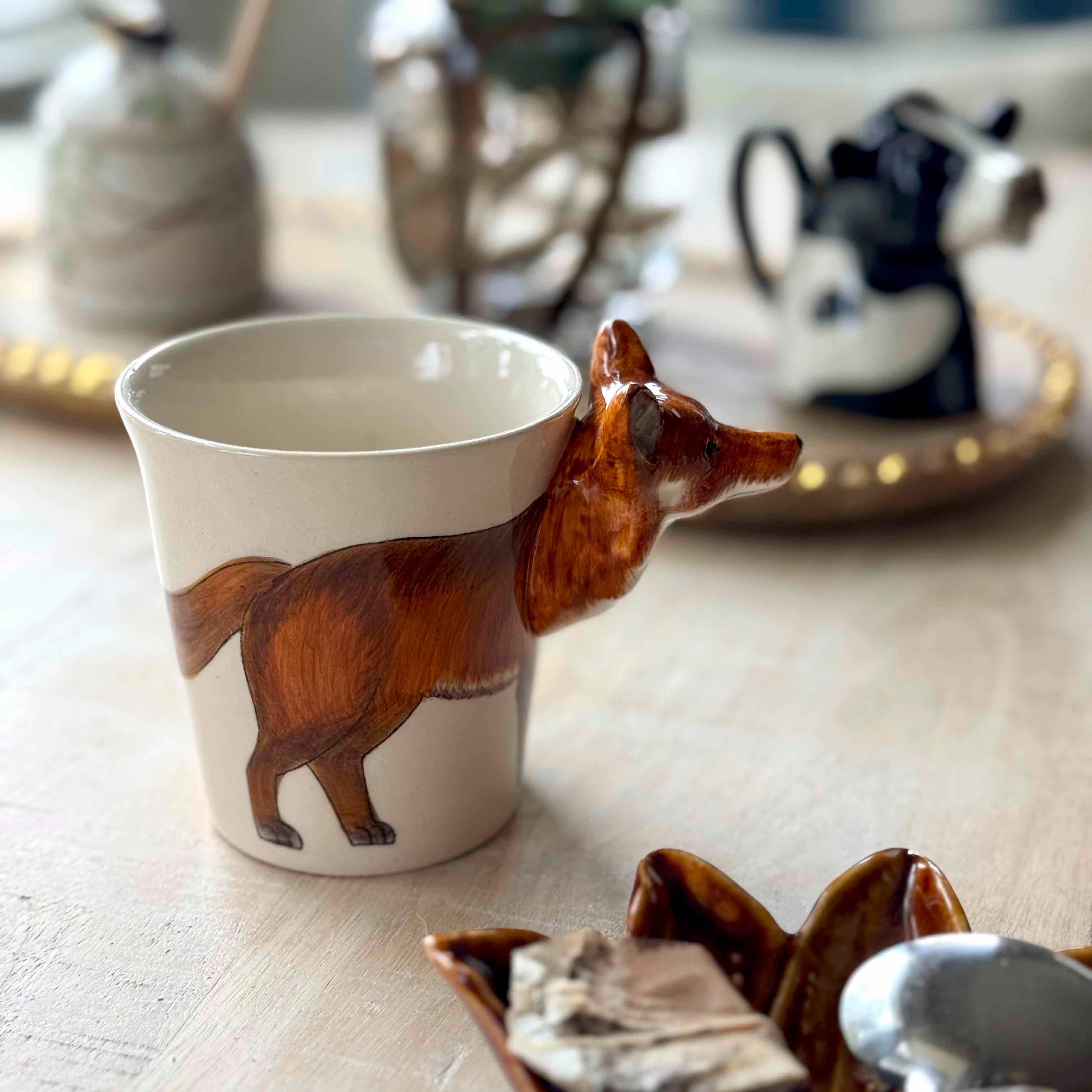 Ceramic mug with a fox design on a wooden surface with decorative items in the background.