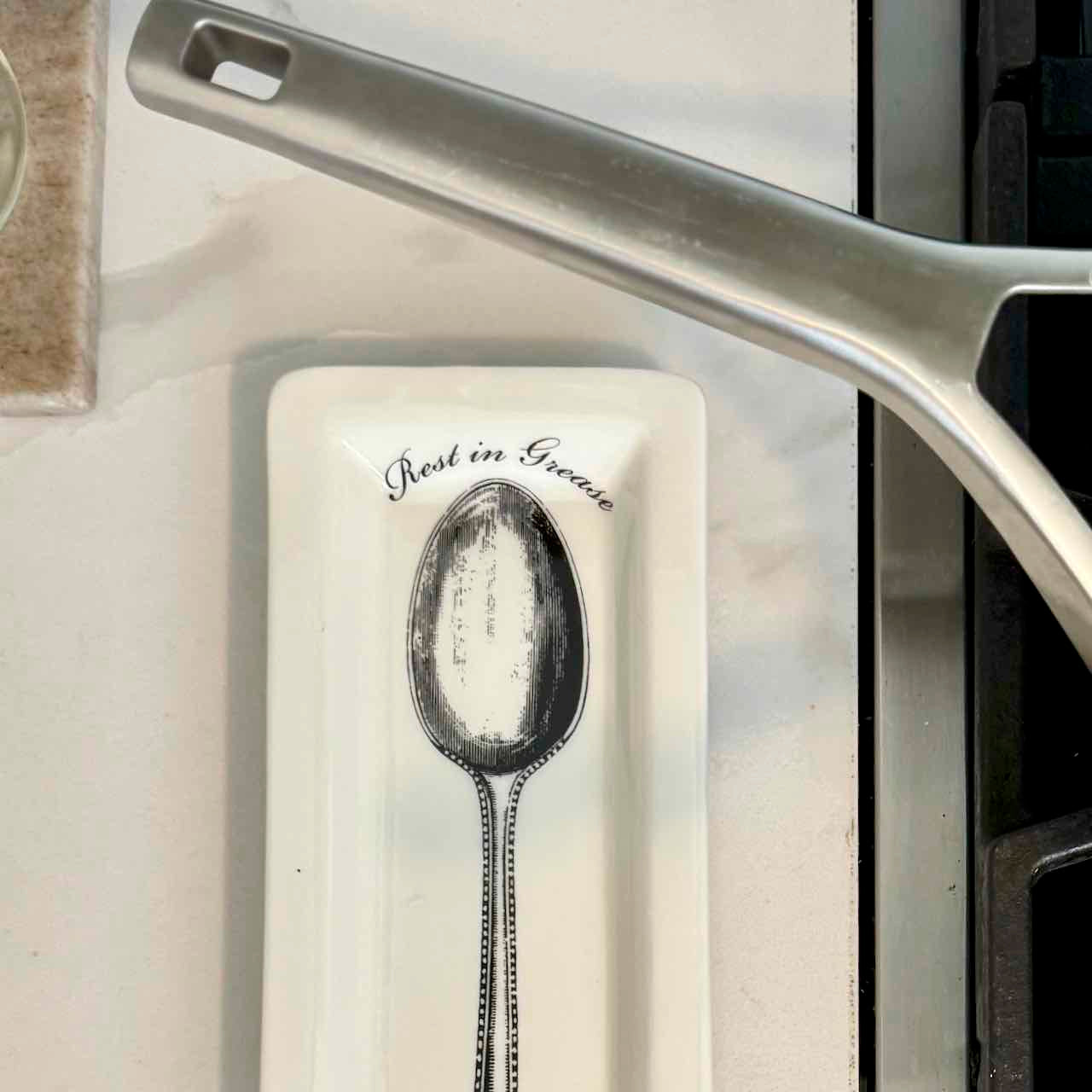 A white ceramic spoon rest with a black illustration of a spoon and the words 'Best in Grease' printed on it, placed on a light-colored countertop next to a stainless steel pan.