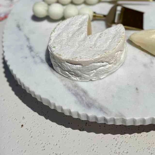 A small wedge of soft white cheese, likely brie or camembert, resting on a white marble serving board with a cheese knife nearby.