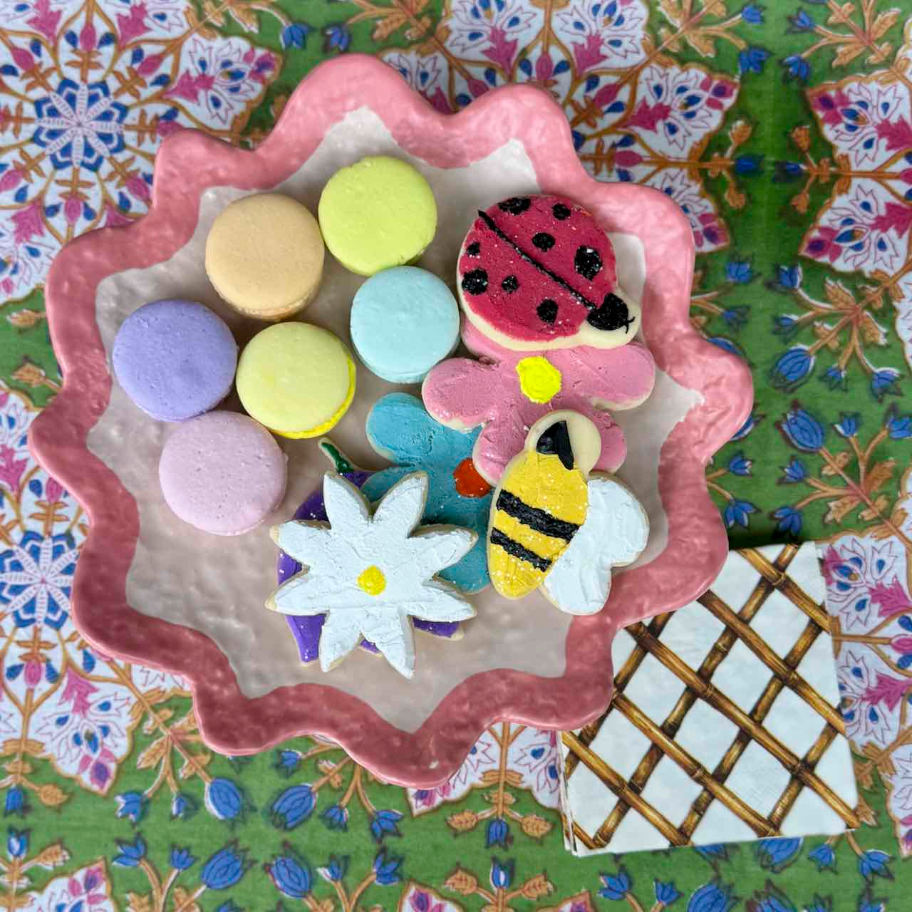 A collection of colorful macarons and decorated cookies arranged on a pink scalloped ceramic plate, set against a green floral fabric background.