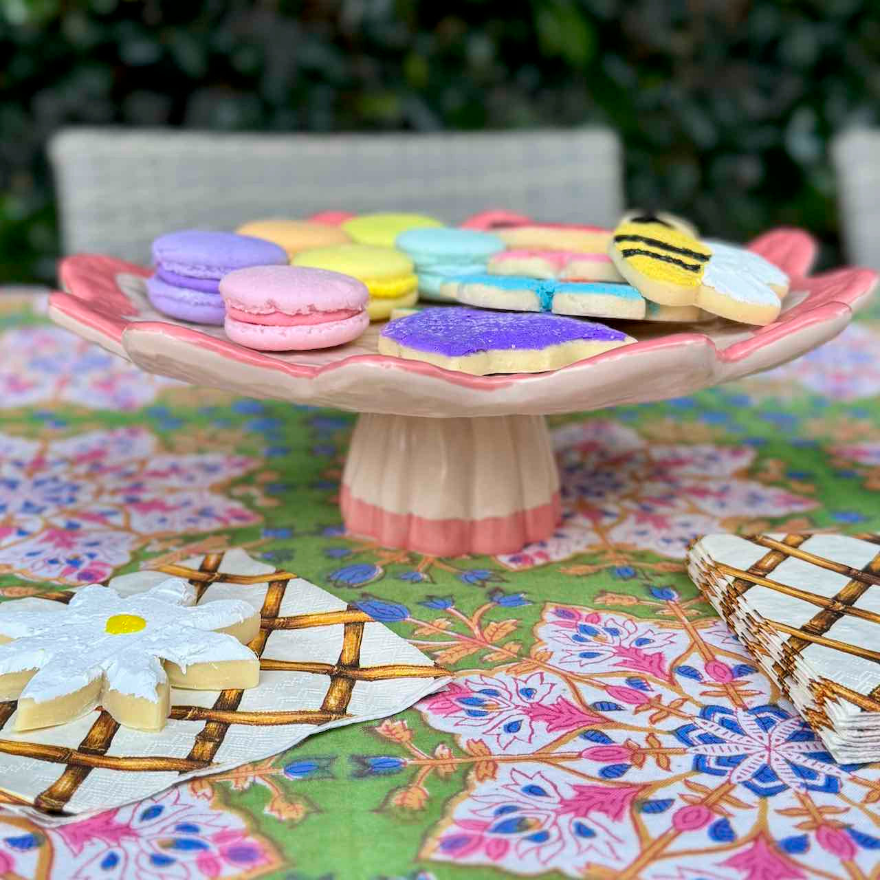 A ceramic flower-shaped cake stand filled with colorful macarons and decorated cookies, set on a floral tablecloth outdoors.
