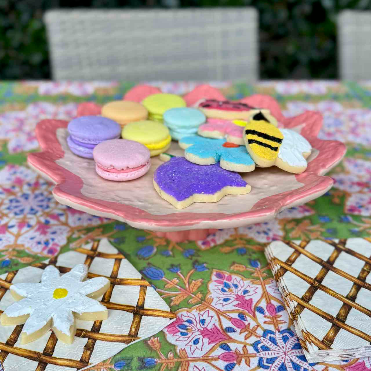 A pink ceramic flower-shaped serving platter holding assorted colorful sugar cookies, including macarons, flower-shaped cookies, and a bee-shaped cookie, set on a floral tablecloth.