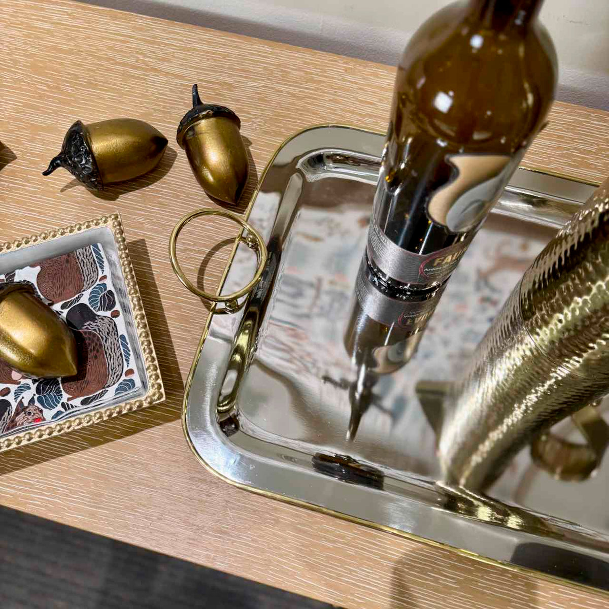 A polished silver rectangular tray on a light wood tabletop, holding a brown glass bottle and a metallic gold snake figurine. Two gold acorn ornaments and a small decorative dish are nearby.