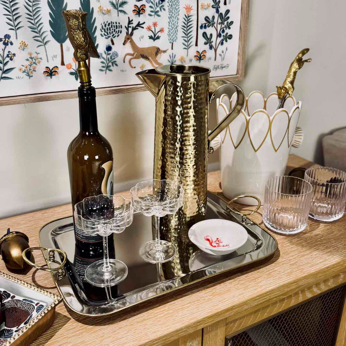 A styled bar setup on a wooden console table featuring a hammered gold metal pitcher, a silver tray, a dark wine bottle with a gold owl decanter, two clear coupe glasses, and a white heart-shaped holder with gold accents.