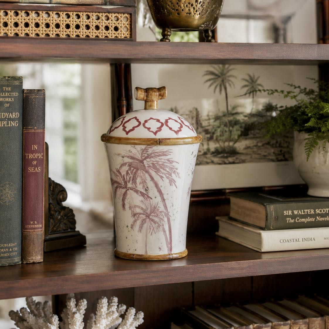 Decorative vase with palm tree design on a wooden shelf with books and plants.