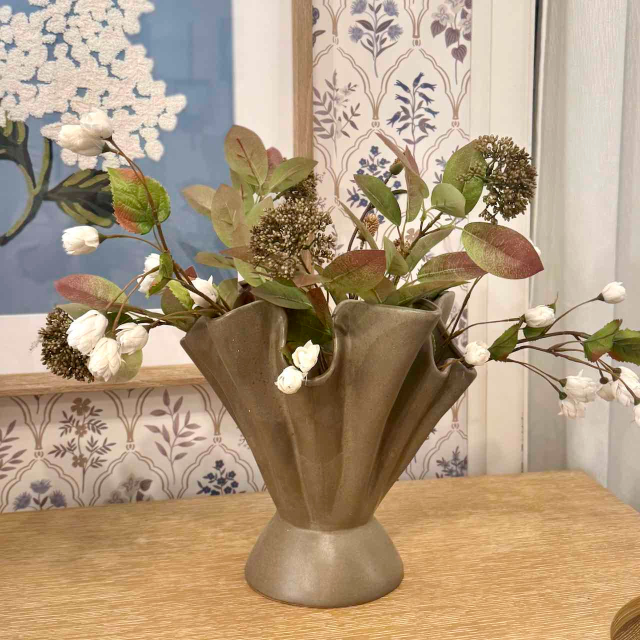 Decorative vase with artificial flowers on a wooden surface with floral wallpaper in the background