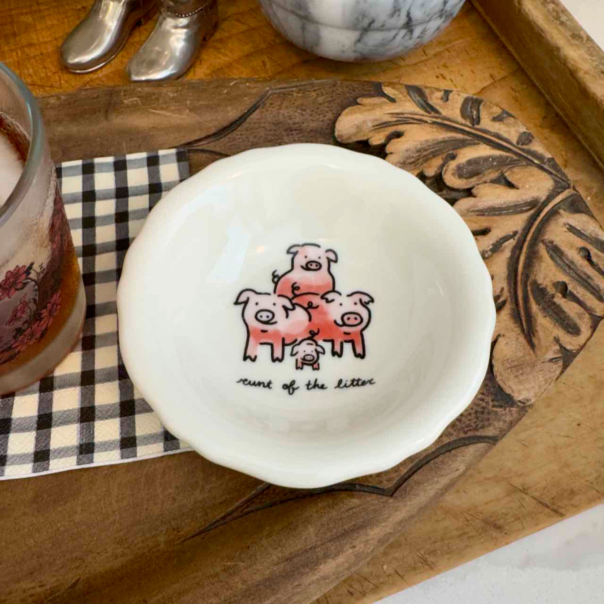 A small white ceramic bowl with a scalloped rim, featuring a hand-drawn illustration of four pink pigs and the text 'count of the litter' inside. The bowl is styled on a carved wooden tray next to a checkered napkin and a glass.