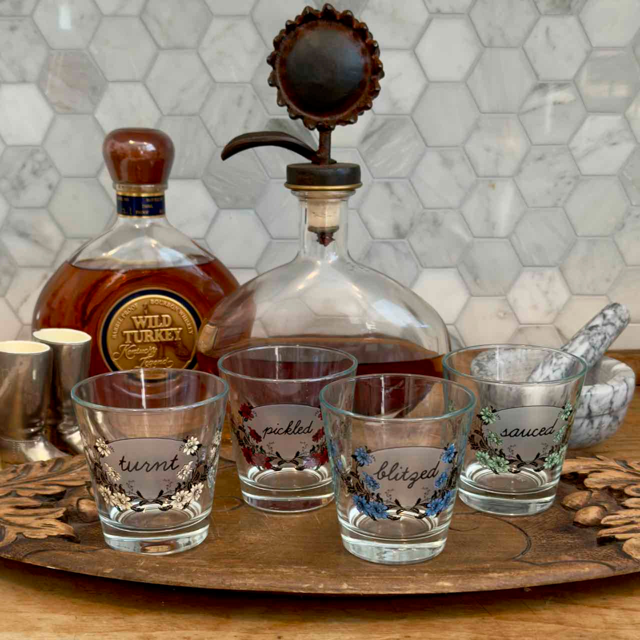 Four clear glass shot glasses with floral designs and handwritten labels reading 'turnt', 'pickled', 'blazed', and 'saucy', arranged on a wooden tray beside a whiskey bottle and decanter in a kitchen setting.