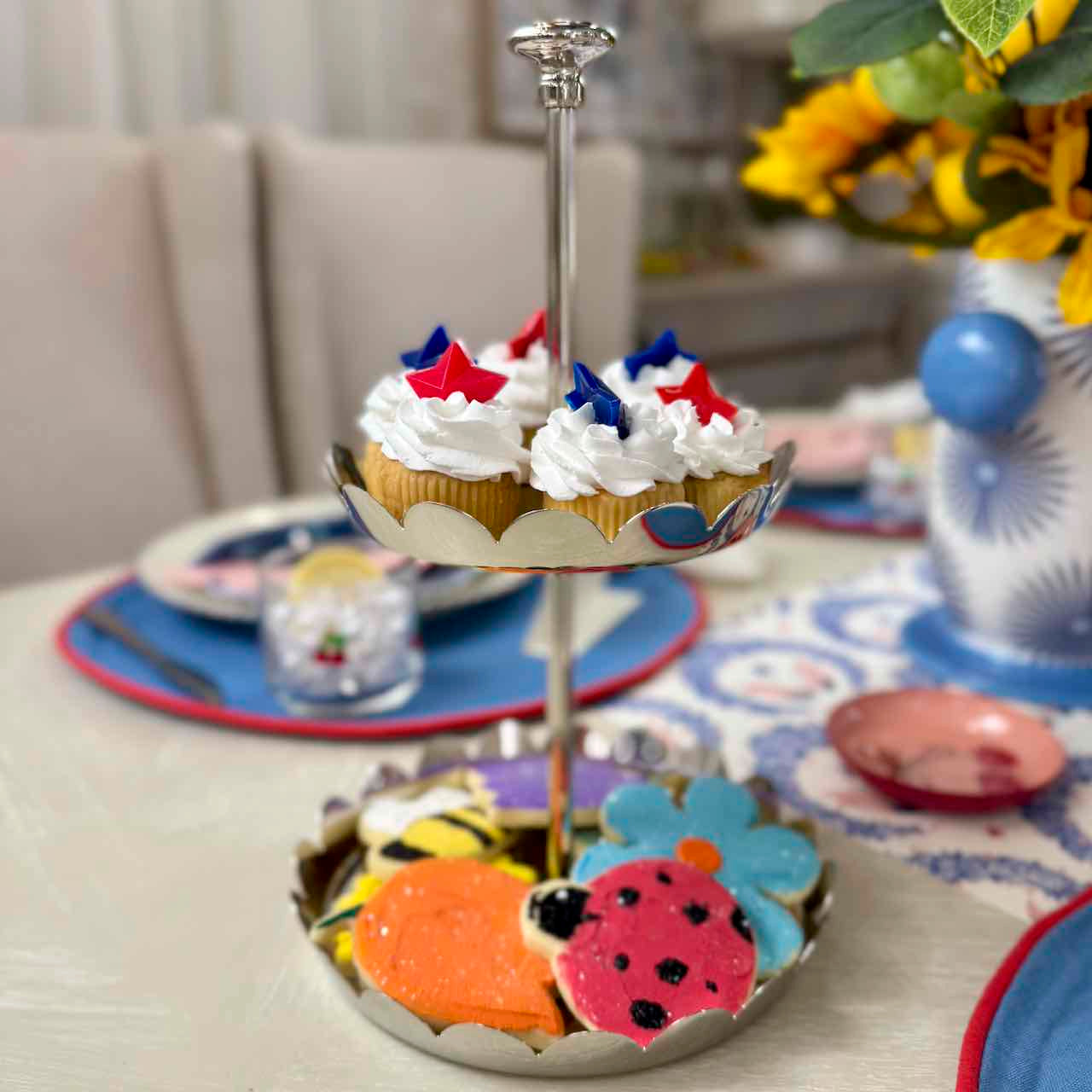 Two cupcakes with red, white, and blue decorations on a two-tiered stand with colorful designs on a table.