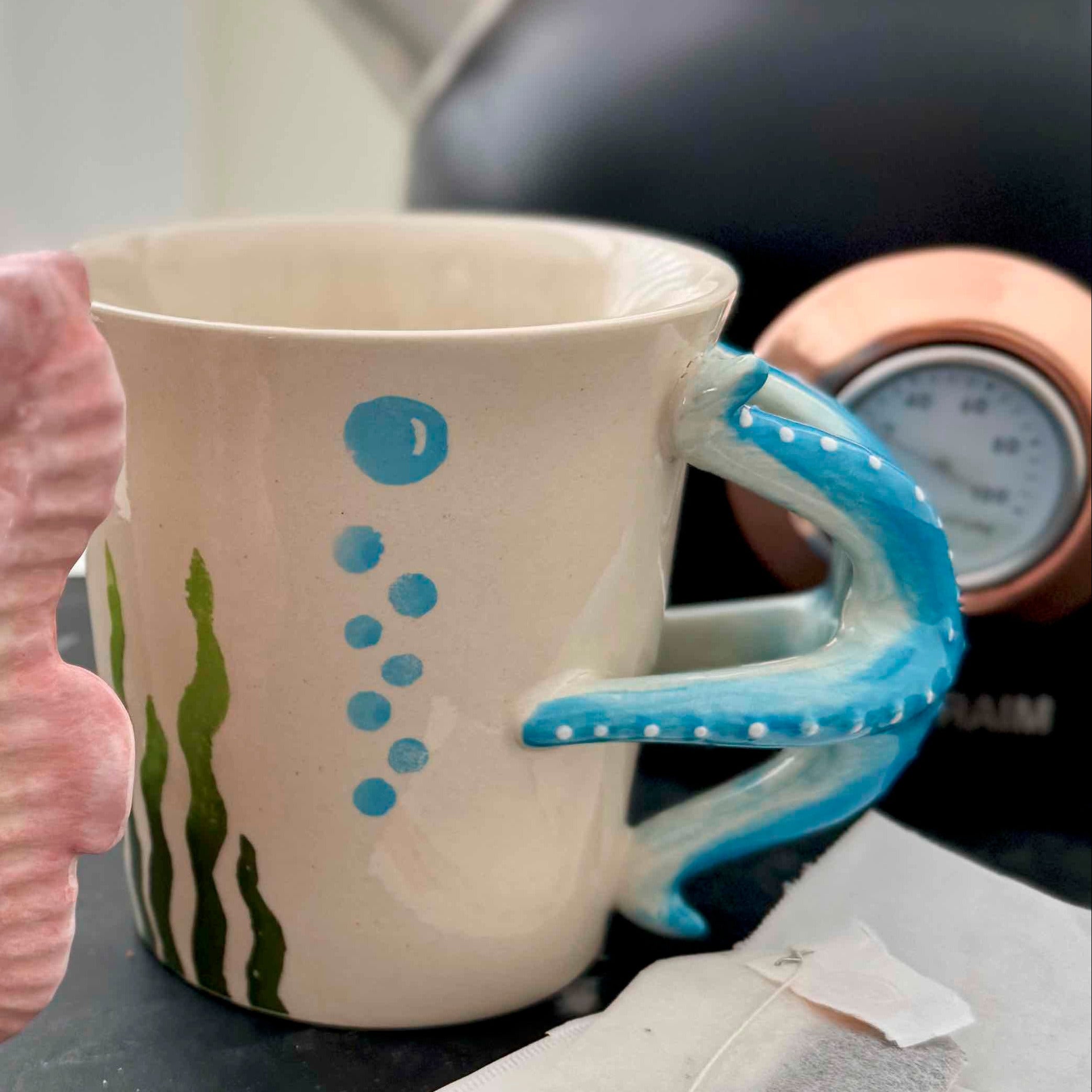 Two ceramic mugs with seahorse handles on a kitchen counter with a teapot and tea bags in the background.