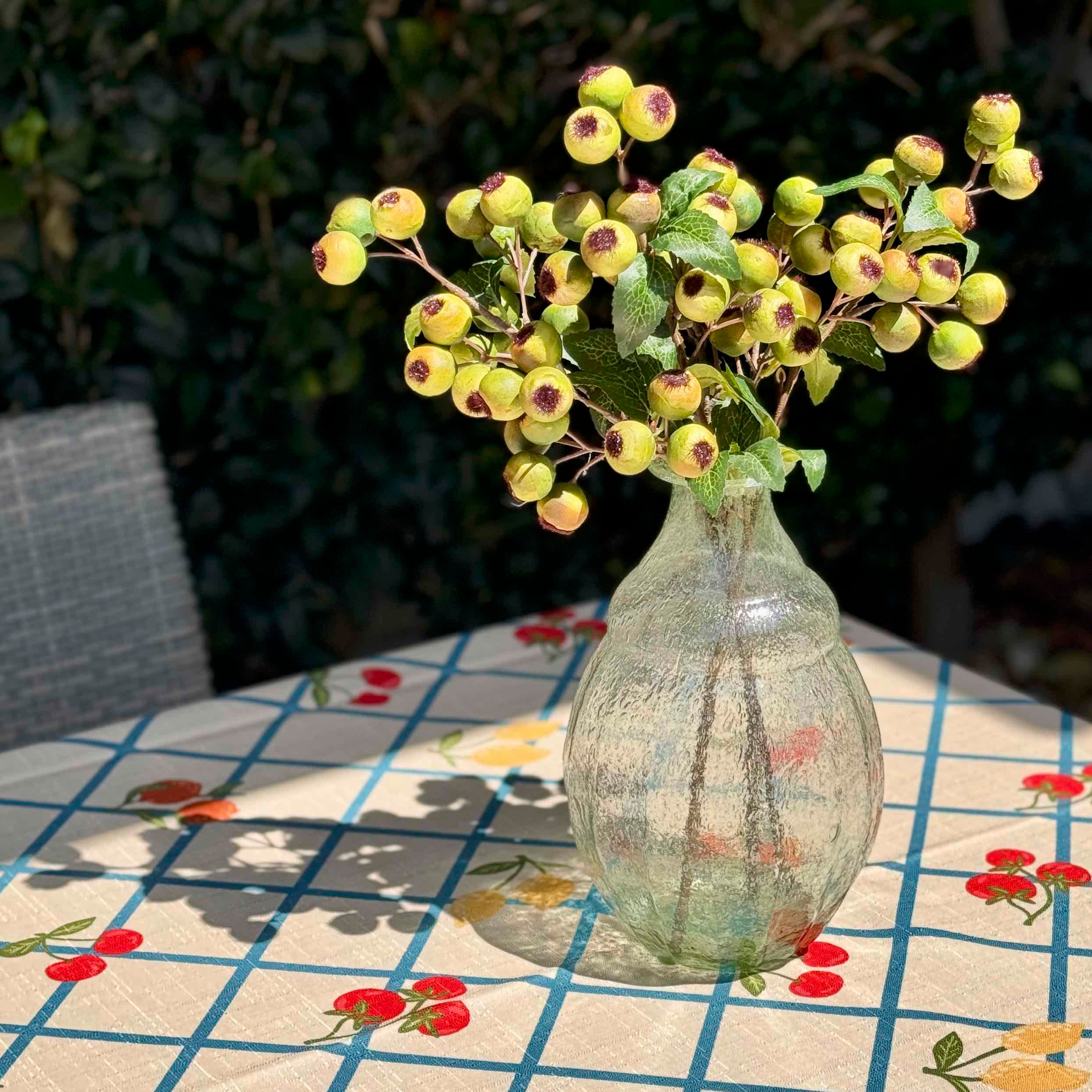 A clear textured glass vase filled with artificial green berries and leaves, placed on a table with a floral tablecloth in an outdoor garden setting.
