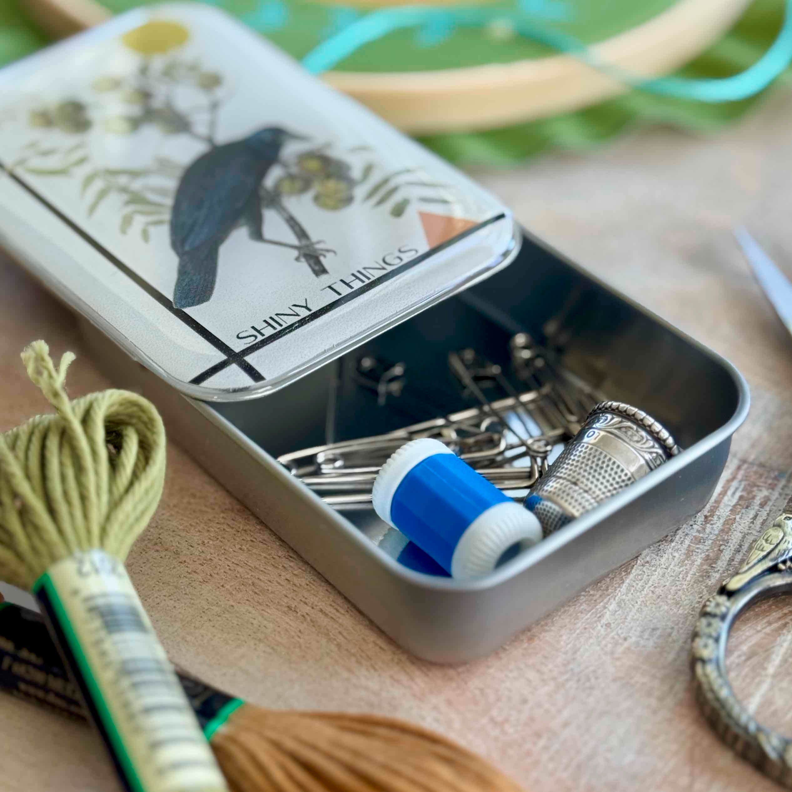 Small metal tin containing sewing supplies with a decorative label on a wooden surface.
