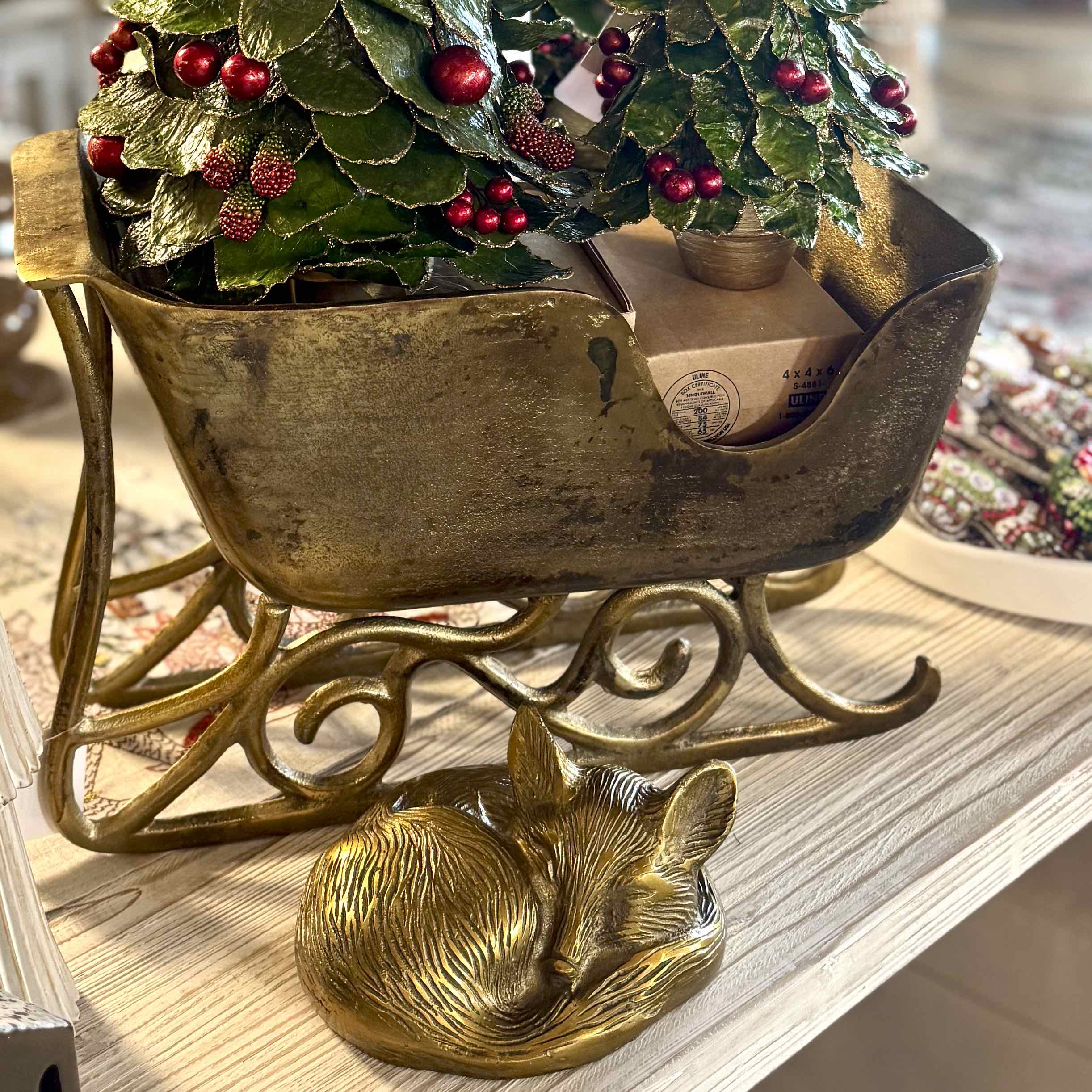 Decorative brass sleigh with a small dog figurine and Christmas tree on a wooden surface.