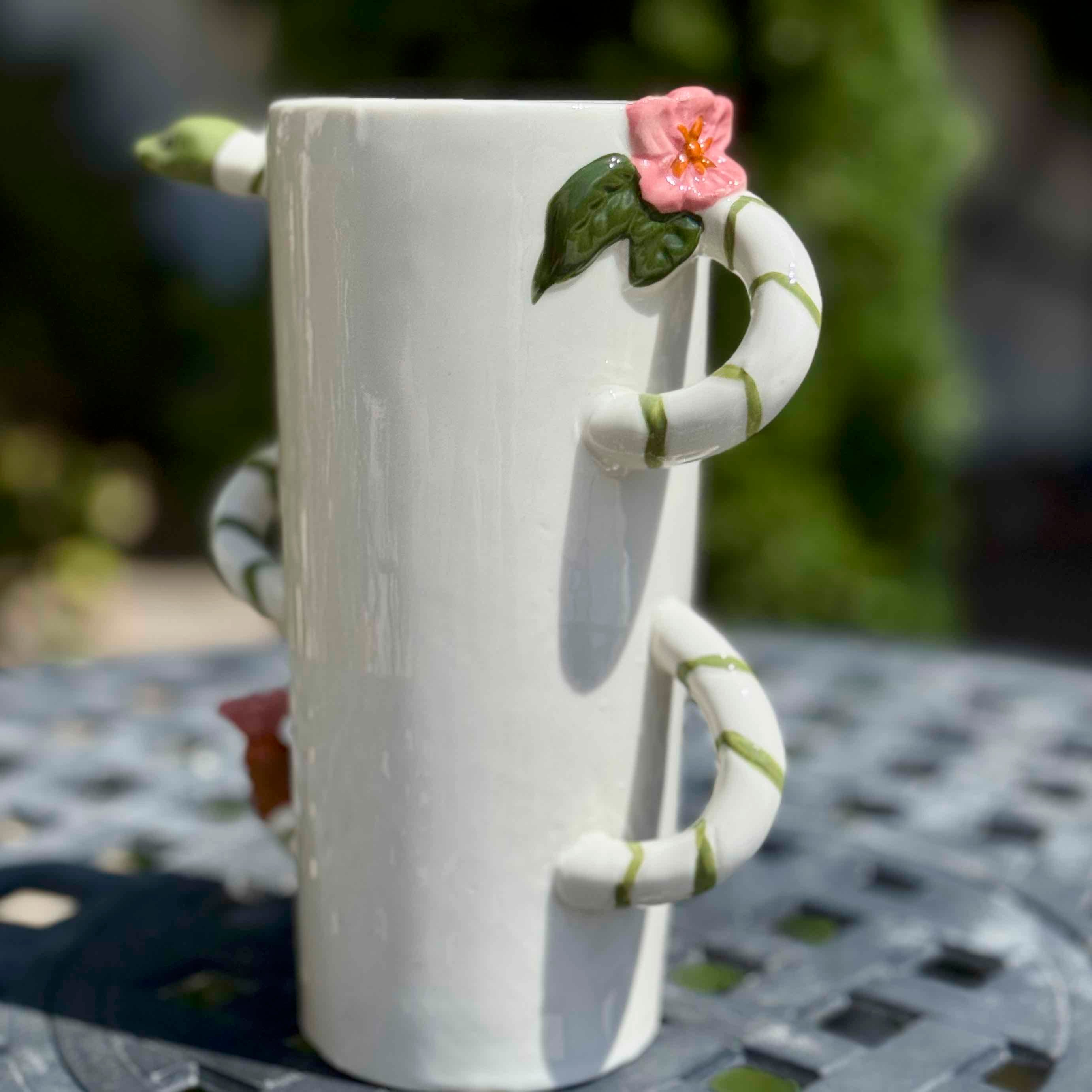 White mug with a decorative handle shaped like a snake and a flower on a metal table outdoors.