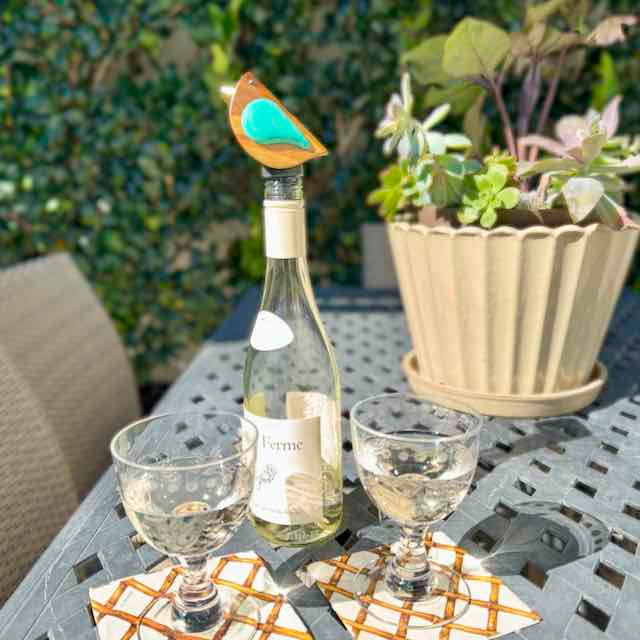 Wine bottle with a decorative bird on top, two glasses, and a potted plant on a table outdoors.