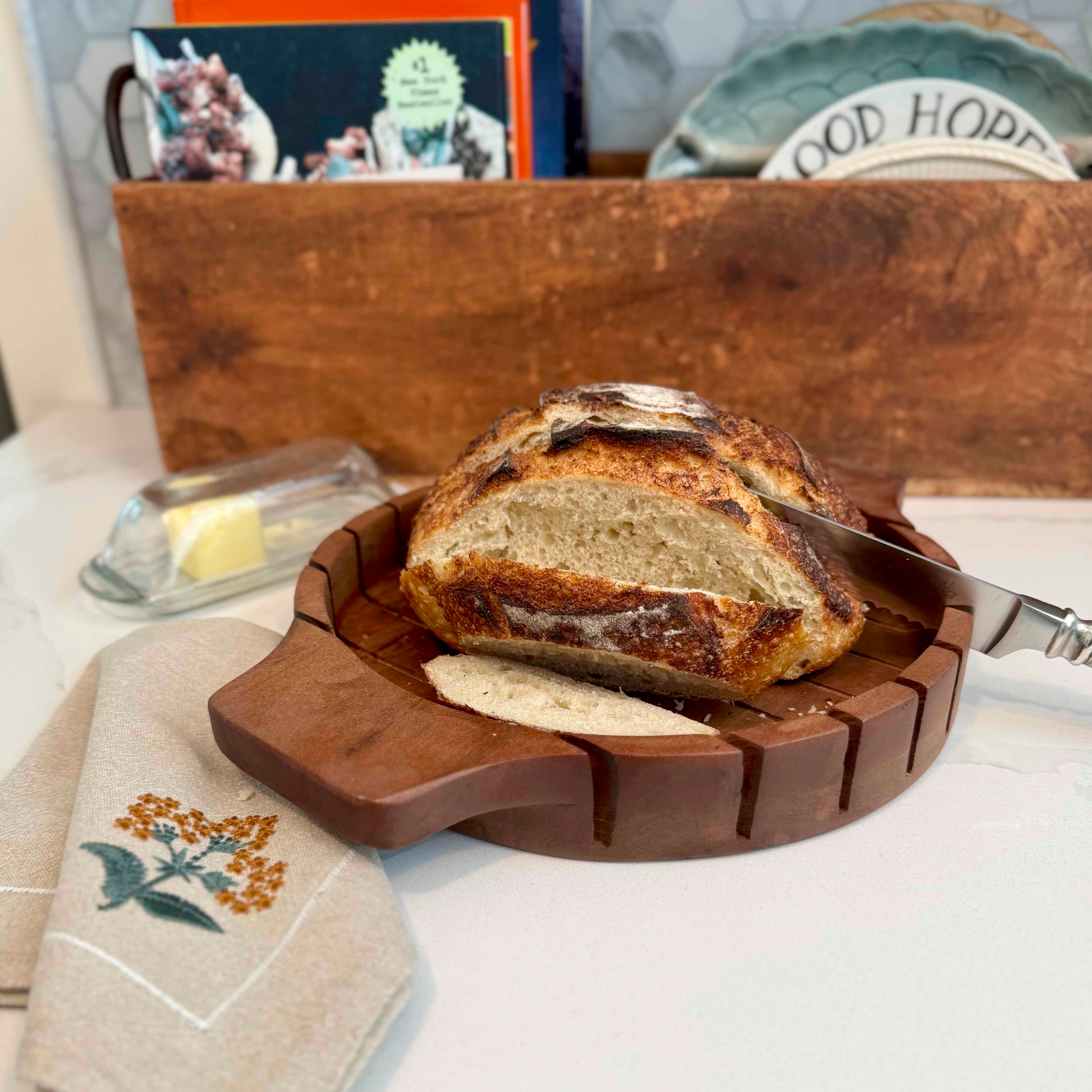 Loaf of bread on a wooden cutting board with a knife, on a table with a book and butter dish in the background.