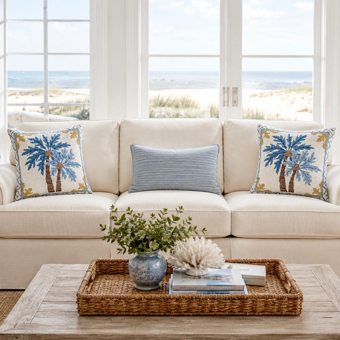A styled living room scene with a cream sofa, two blue-and-gold palm tree throw pillows, and a light blue striped accent pillow in front of large windows overlooking a beach. A woven tray on a wooden table holds a small plant and decorative items.