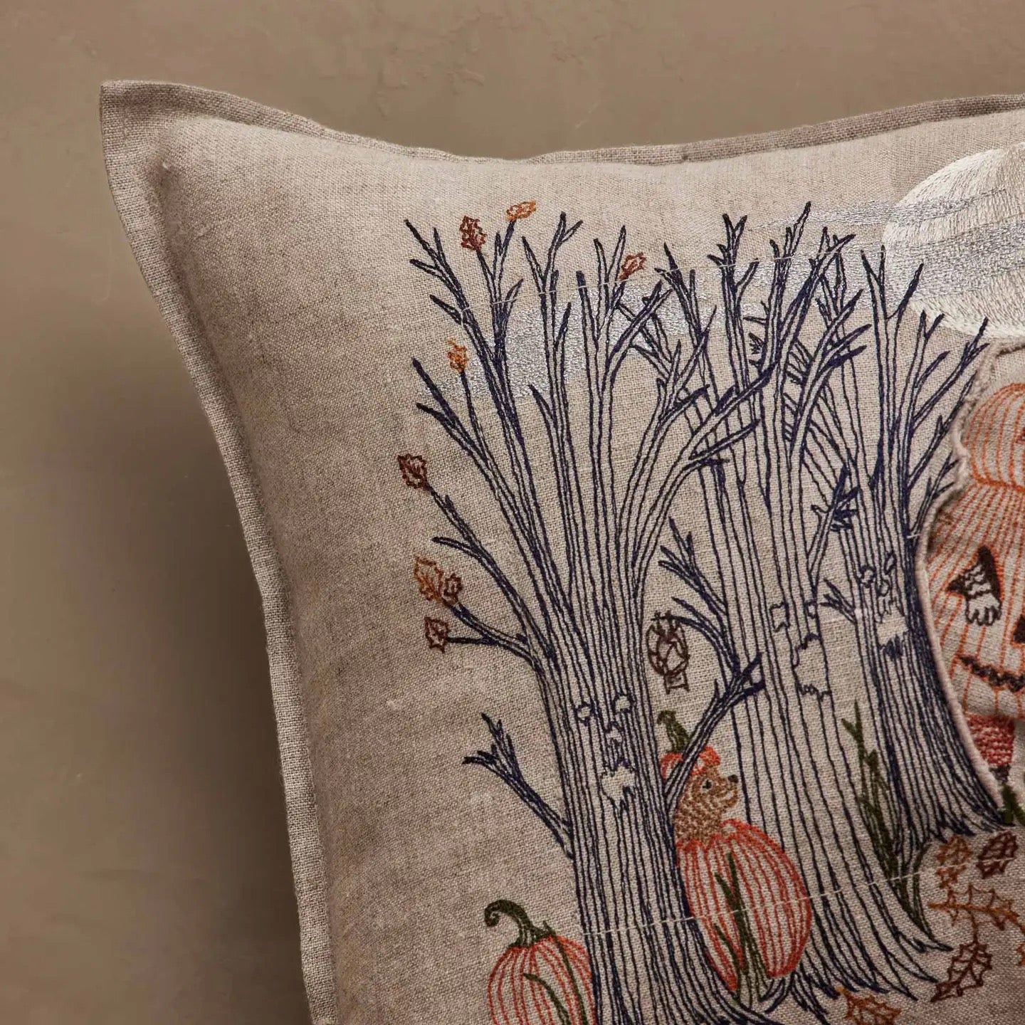 A close-up of a square linen throw pillow featuring a detailed embroidered design of bare trees, acorns, and orange pumpkins on a warm beige background.