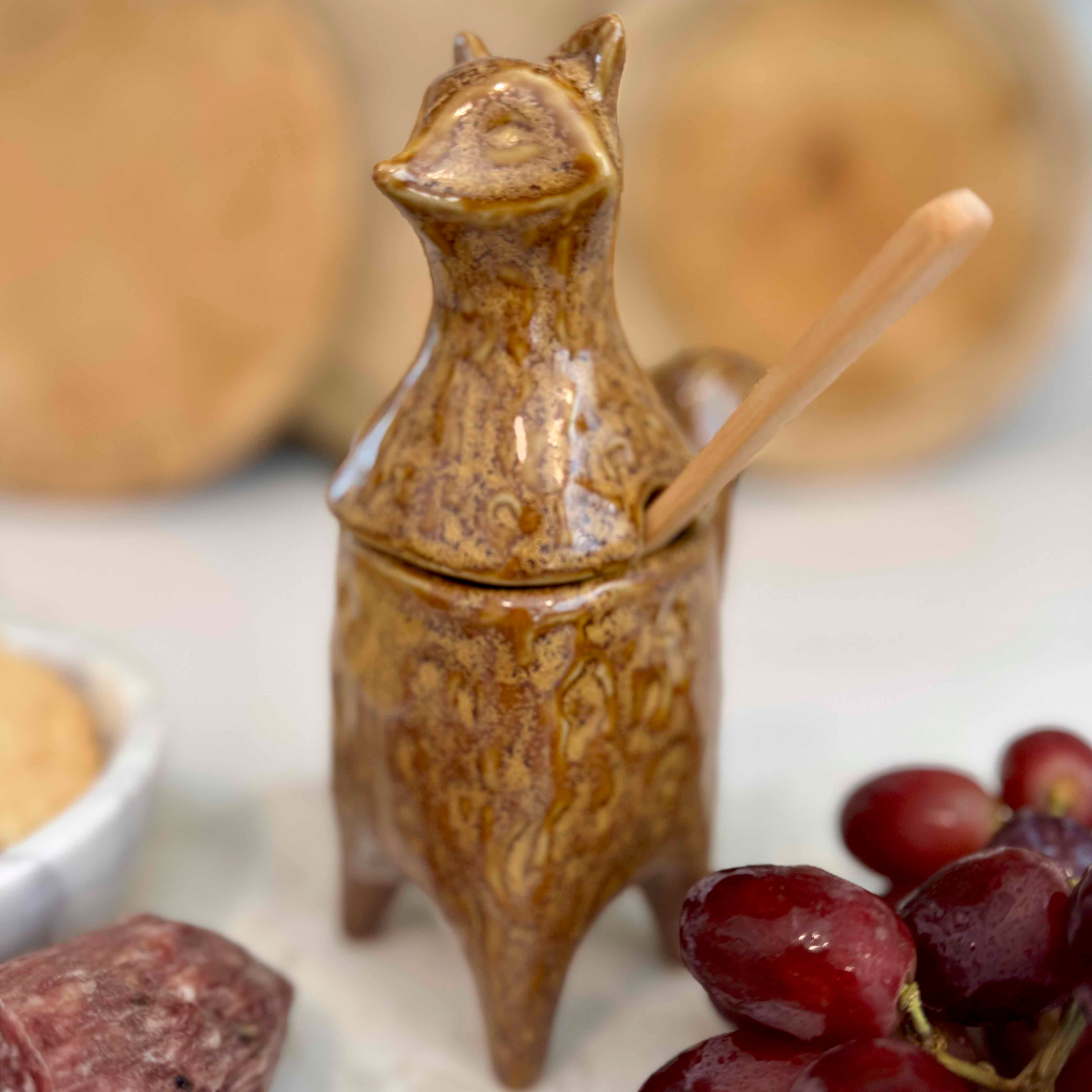 Ceramic fox-shaped container with a wooden spoon on a white surface with grapes and honey.